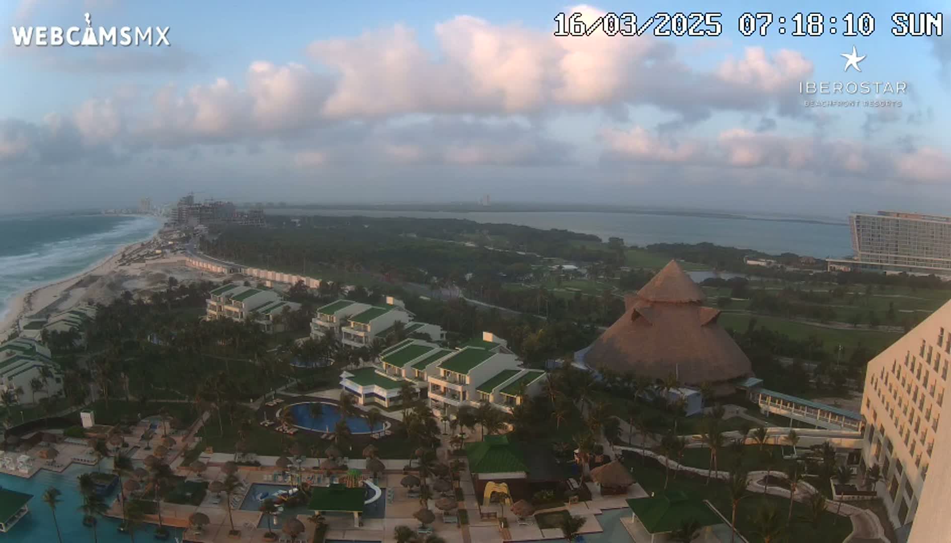An aerial view of a resort complex on a partly cloudy morning, showing buildings, pools, landscaping, and a beach with ocean waves.