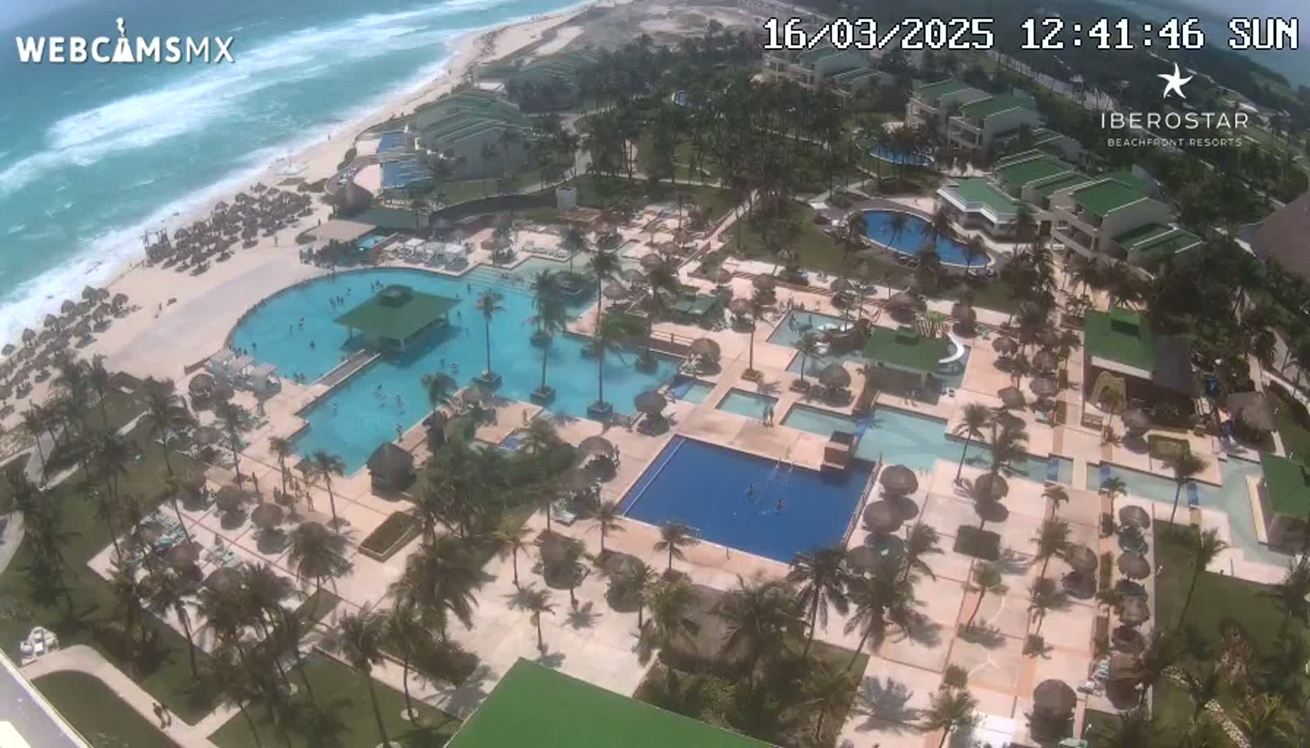 An aerial view shows a resort with multiple pools, palm trees, and a beach with wavy ocean under sunny skies.