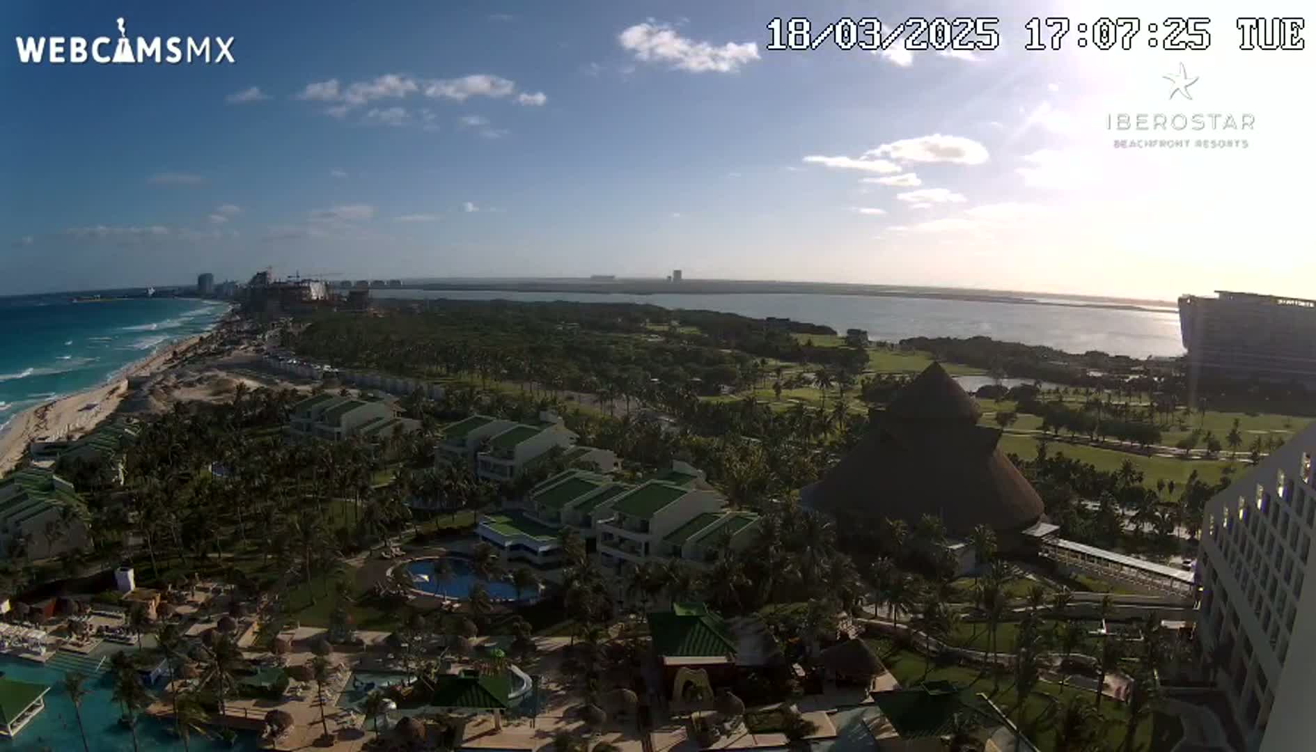 An aerial view of a resort area on a partly sunny day, showing a beach, ocean, and numerous buildings surrounded by lush vegetation.