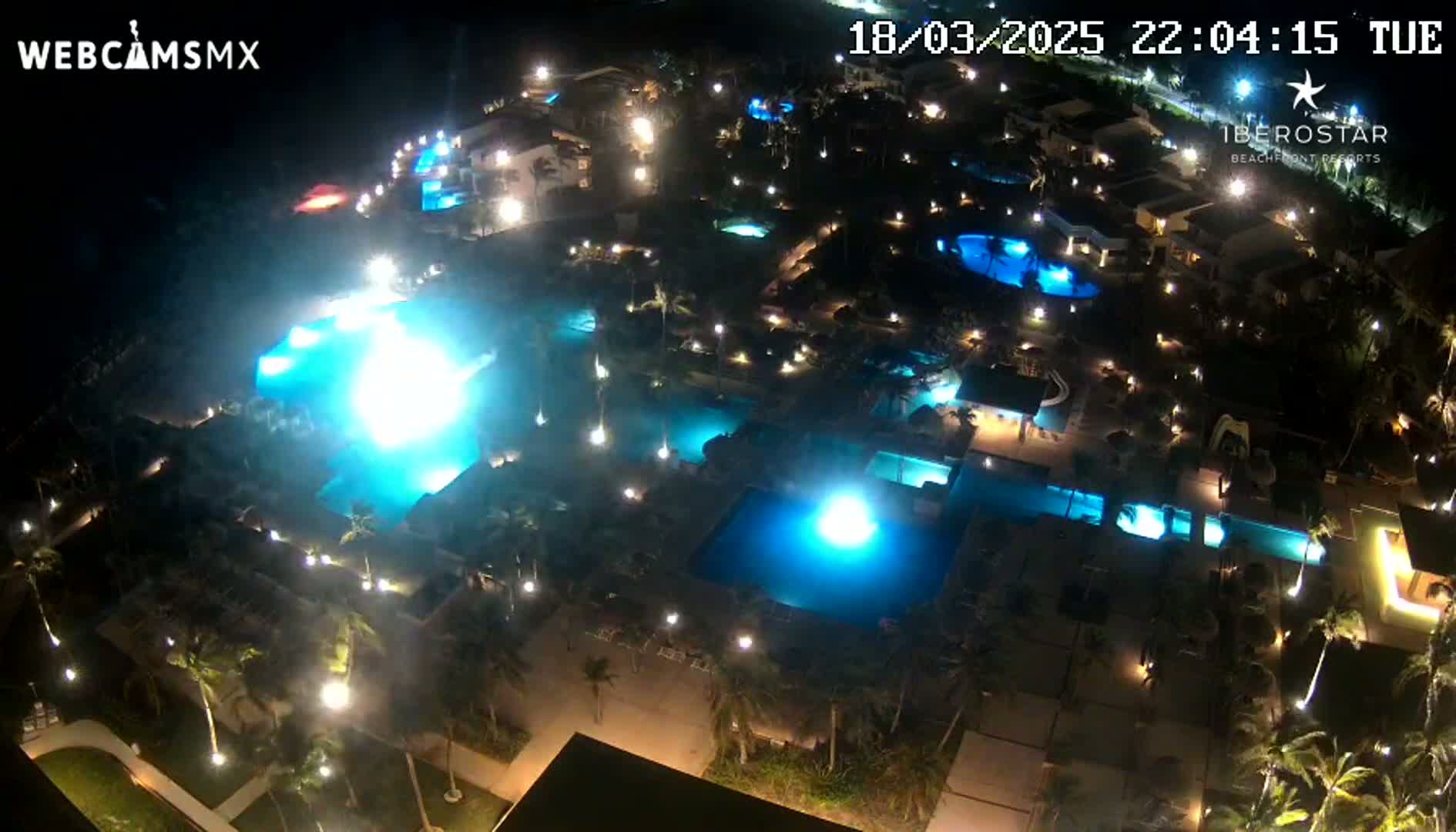 An aerial nighttime view shows a resort complex with numerous illuminated swimming pools and surrounding landscaping.