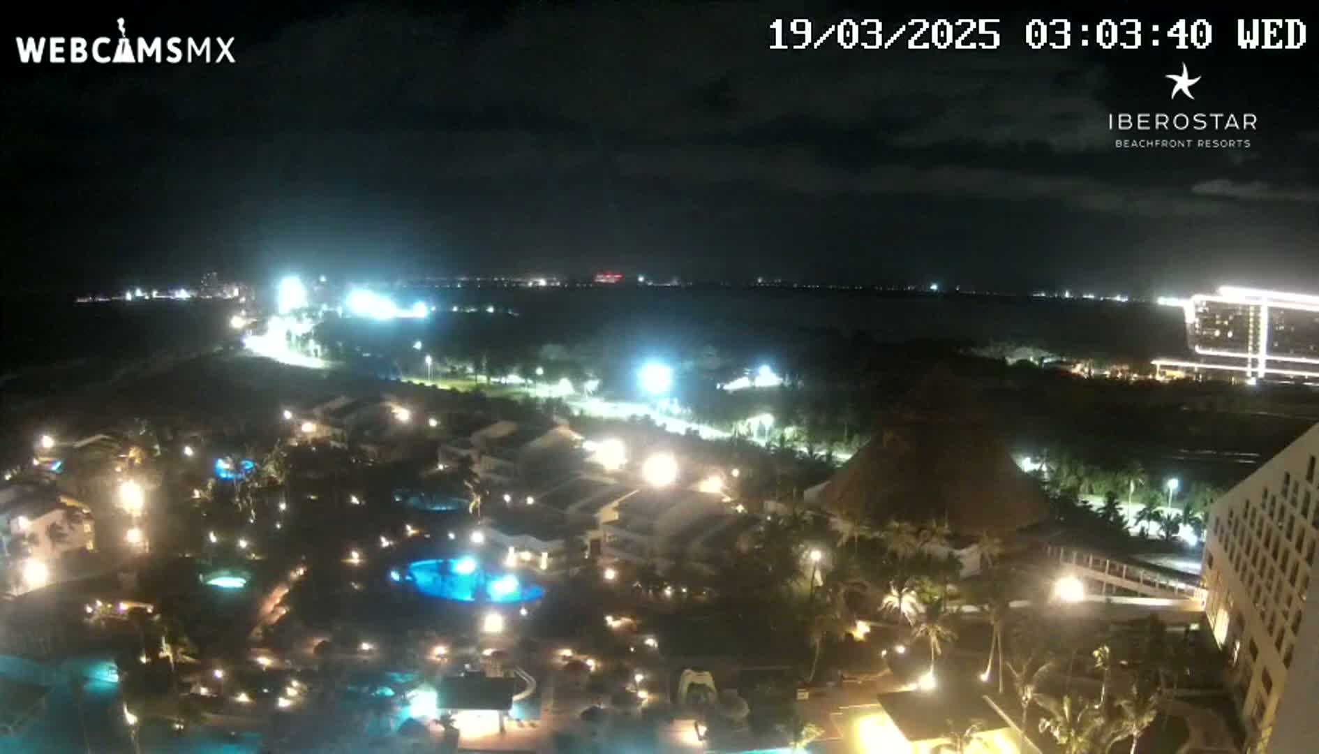 A nighttime aerial view shows a resort complex with illuminated pools and buildings, extending to a dark ocean under a mostly clear sky.