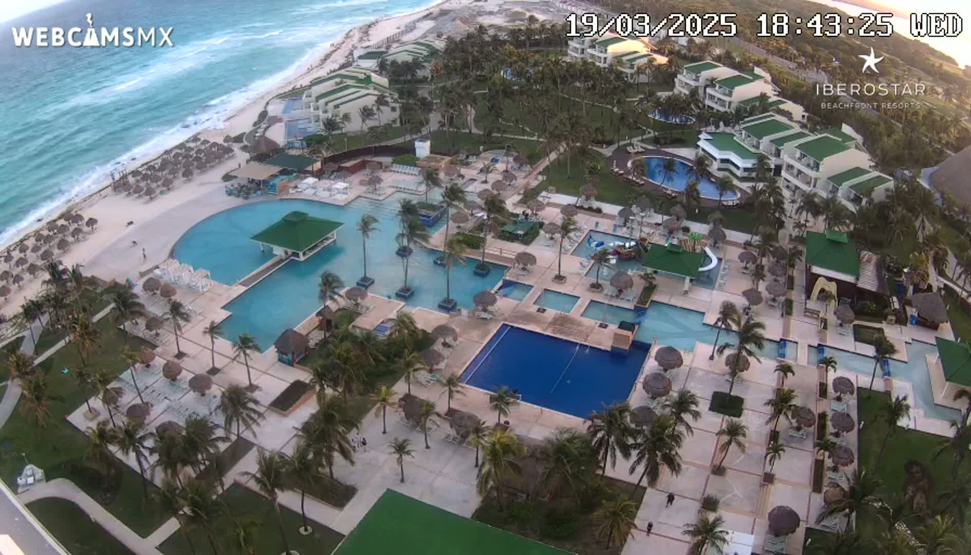 An aerial view of a resort complex with multiple pools and palm trees, situated on a beach with a wavy ocean under a partly cloudy sunset sky.