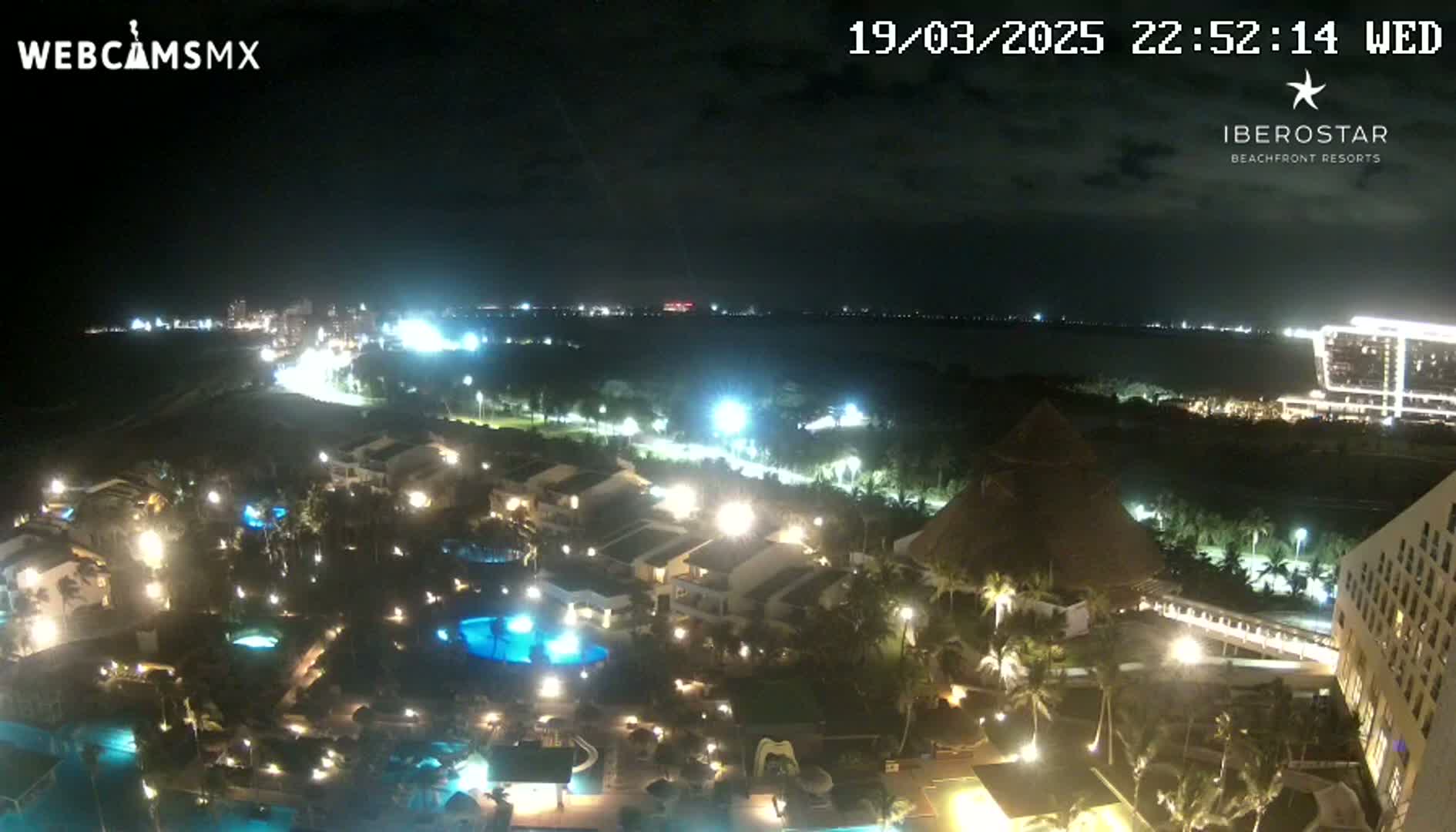 A nighttime, aerial view of a resort area, showing illuminated buildings, pools, and landscaping next to a dark ocean under a partly cloudy sky.