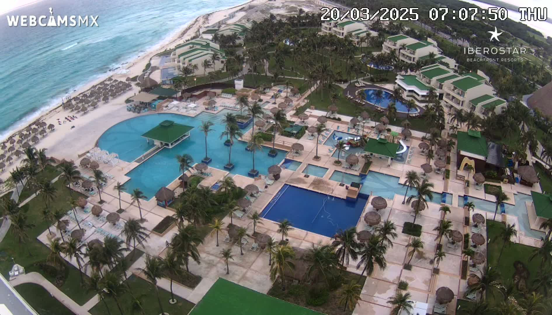 An aerial view shows a resort with multiple pools, a beach, and palm trees under partly cloudy skies.