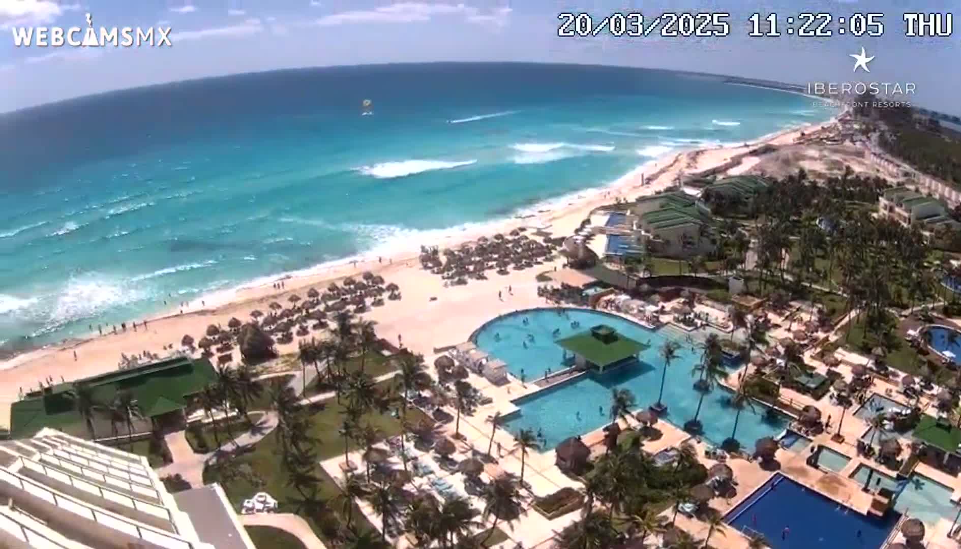 An aerial view shows a resort with multiple pools and a beach on a sunny day with light waves in the turquoise ocean.