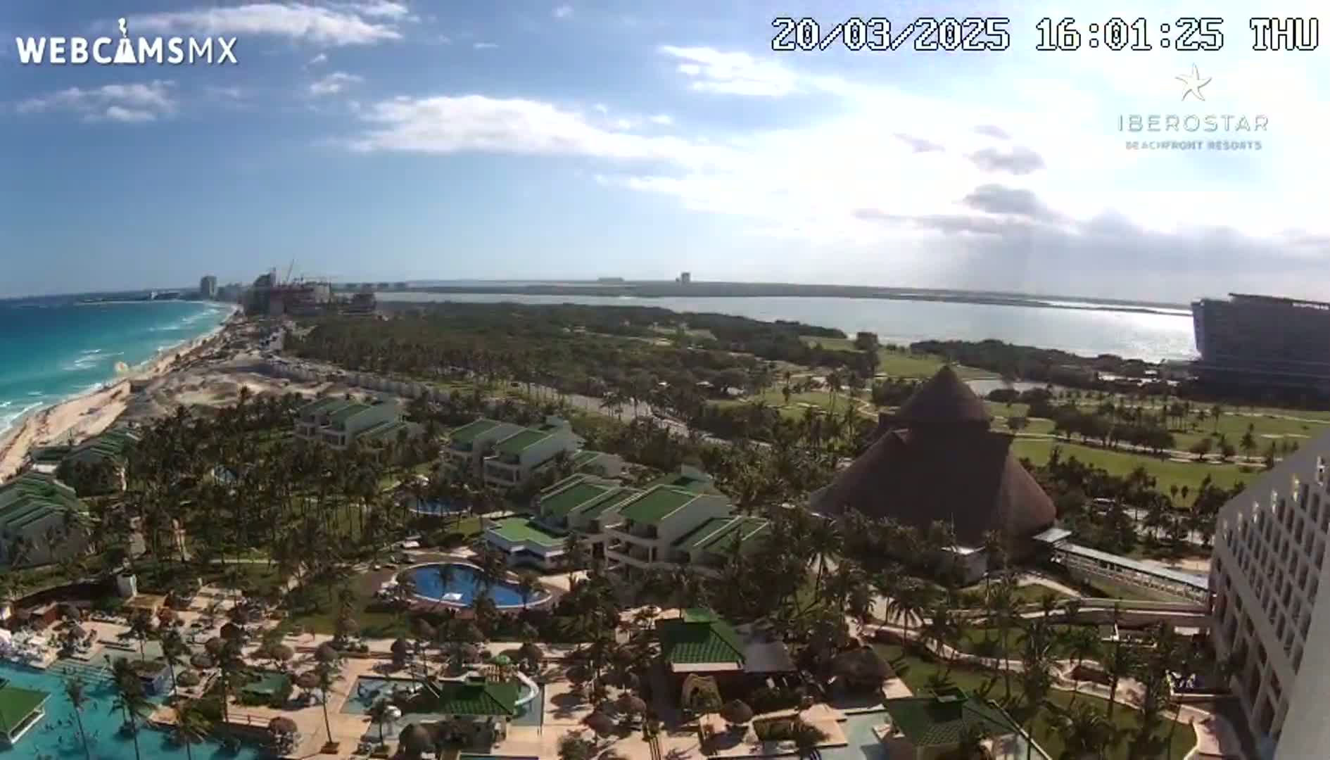An aerial view shows a resort complex with pools and palm trees next to a beach and ocean under a mostly sunny sky.