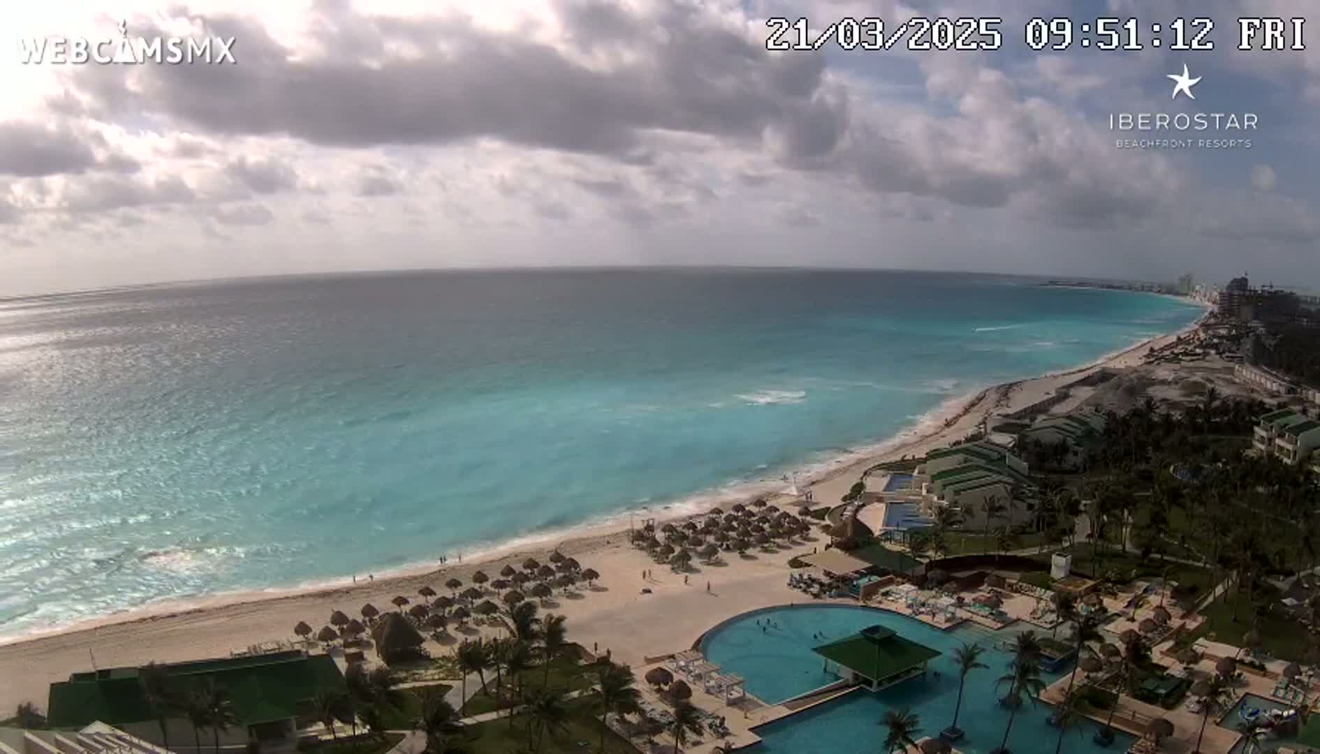 An aerial view shows a beach resort with pools next to a turquoise ocean under a partly cloudy sky.
