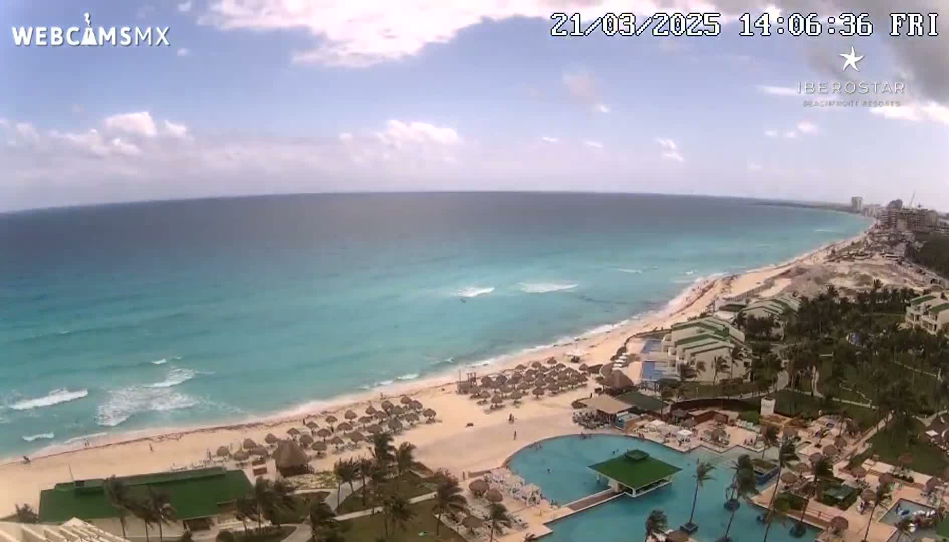 A partly cloudy day shows a beach resort with a large swimming pool complex next to a sandy beach and turquoise ocean with small waves.