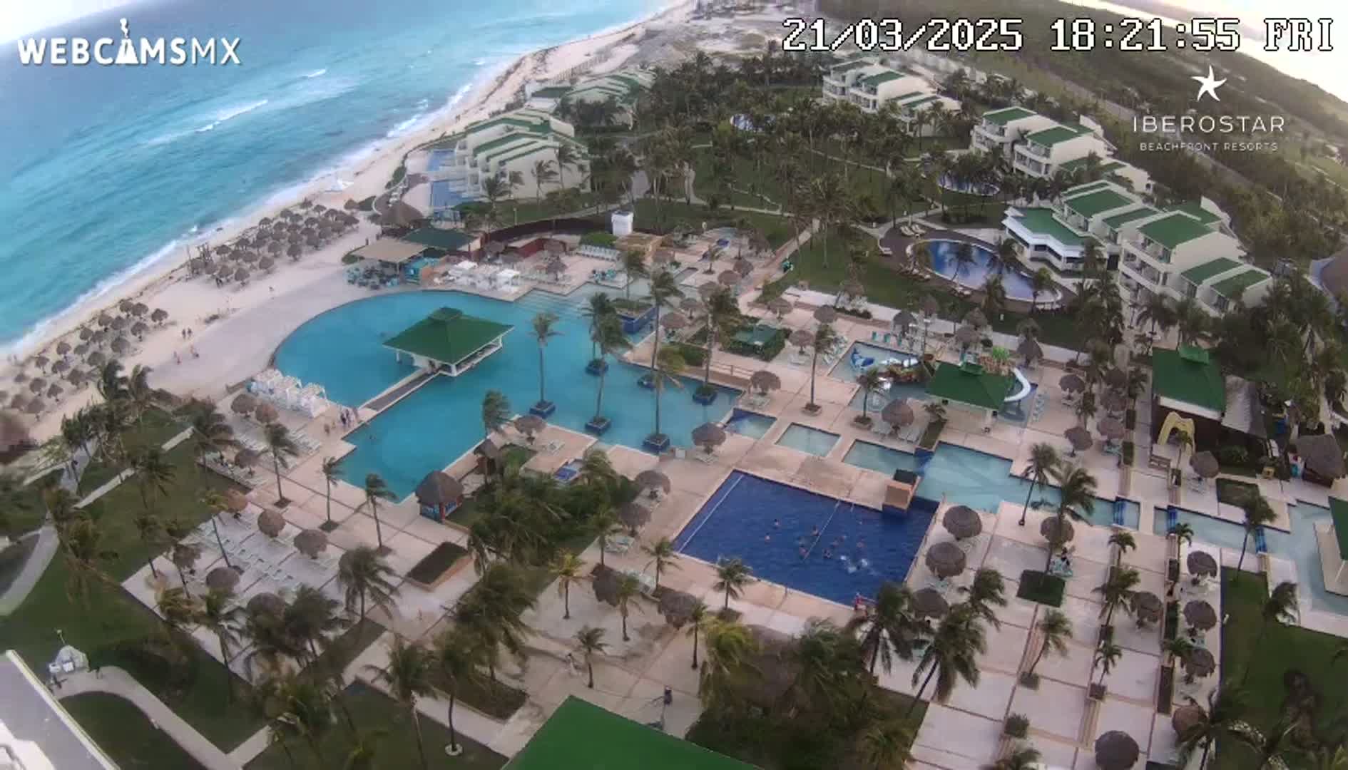 An aerial view of a beachfront resort with multiple pools and palm trees, under windy conditions.