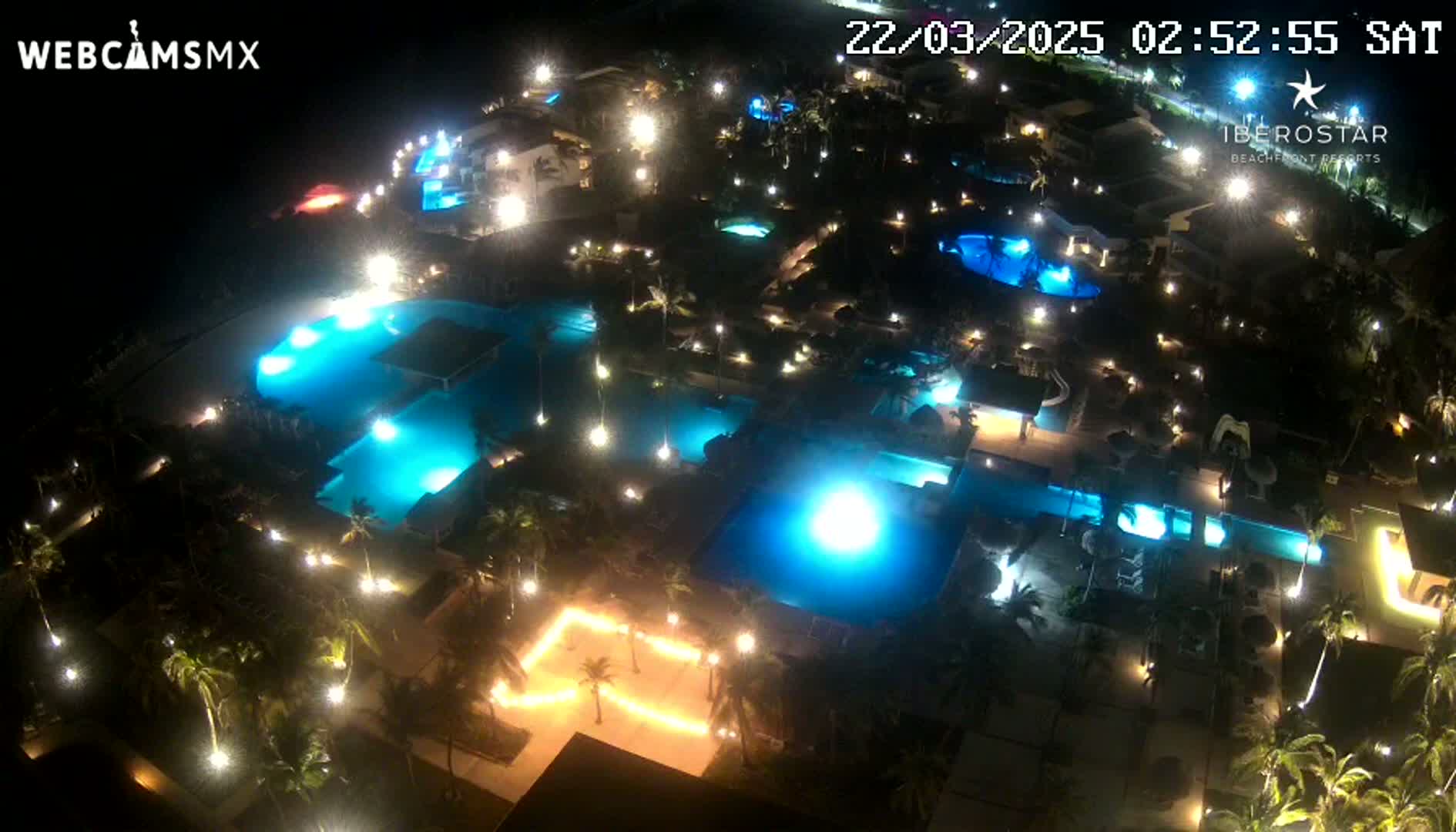 An aerial nighttime view of a resort complex with numerous illuminated swimming pools surrounded by palm trees and buildings.