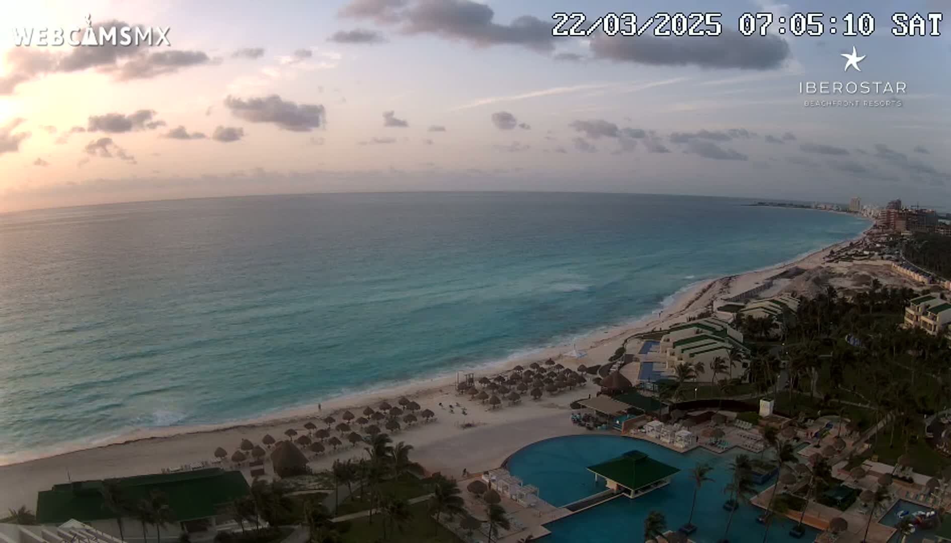 A tranquil sunrise over a calm turquoise ocean and sandy beach, showcasing a resort with pools and beach umbrellas.