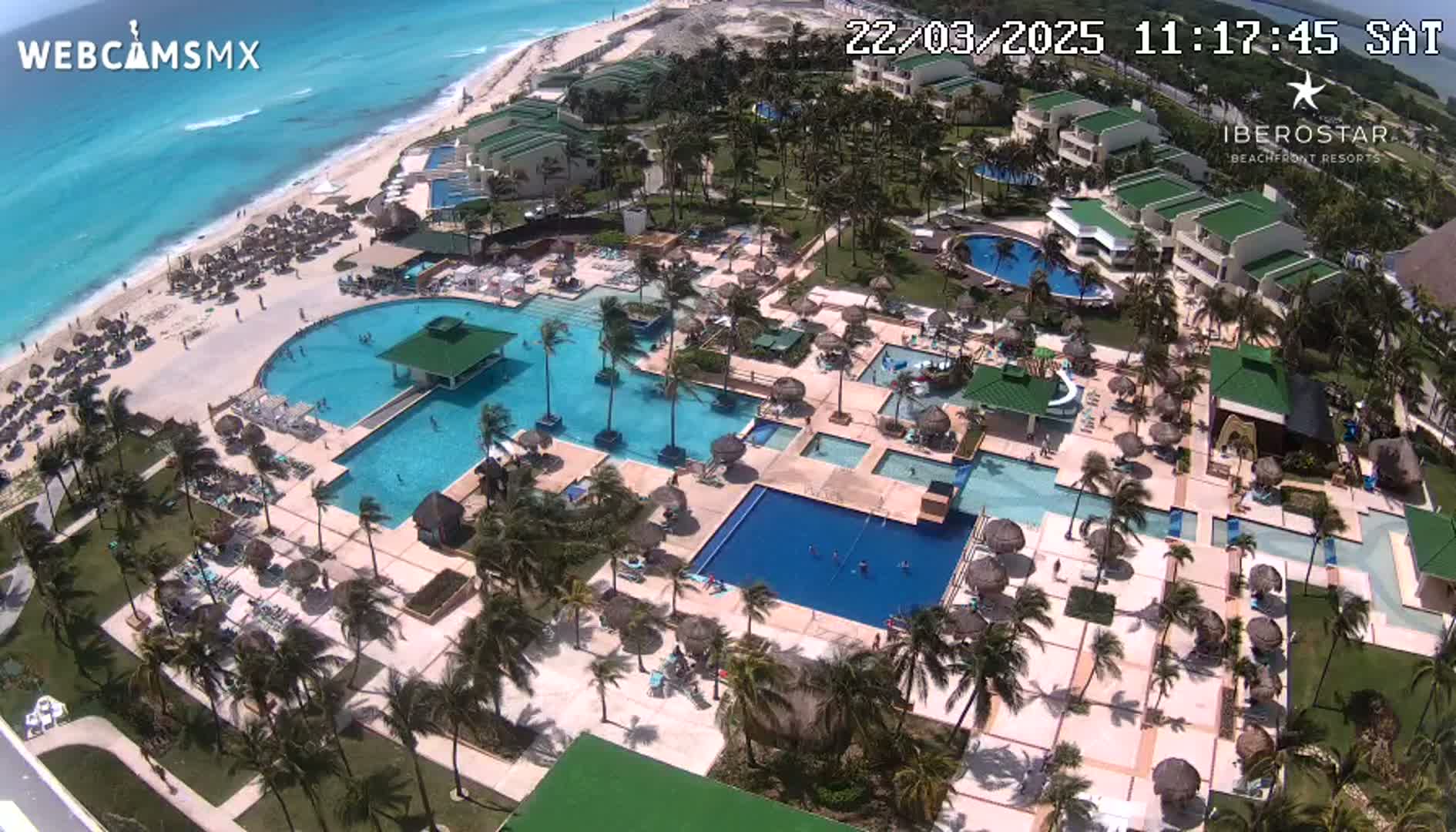 An aerial view of a resort with multiple swimming pools, a beach, and palm trees on a sunny day.
