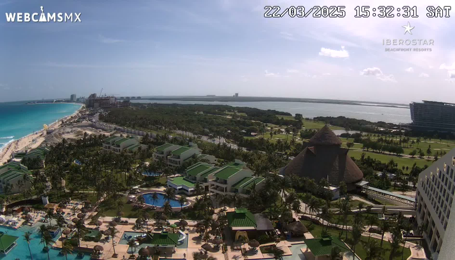An aerial view of a resort area on a sunny day, showing turquoise ocean waters, a beach, lush green landscaping, low-rise buildings with swimming pools, and a larger hotel in the background.