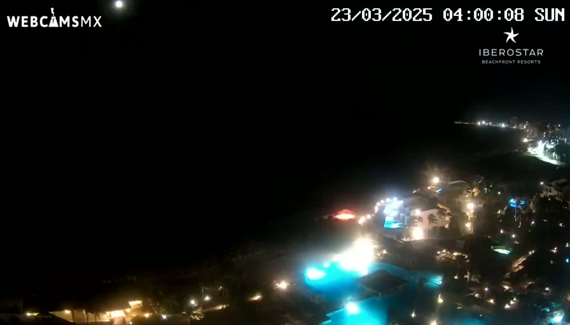 A nighttime aerial view shows a resort complex illuminated by lights, with a bright moon visible in the dark sky above a body of dark water.