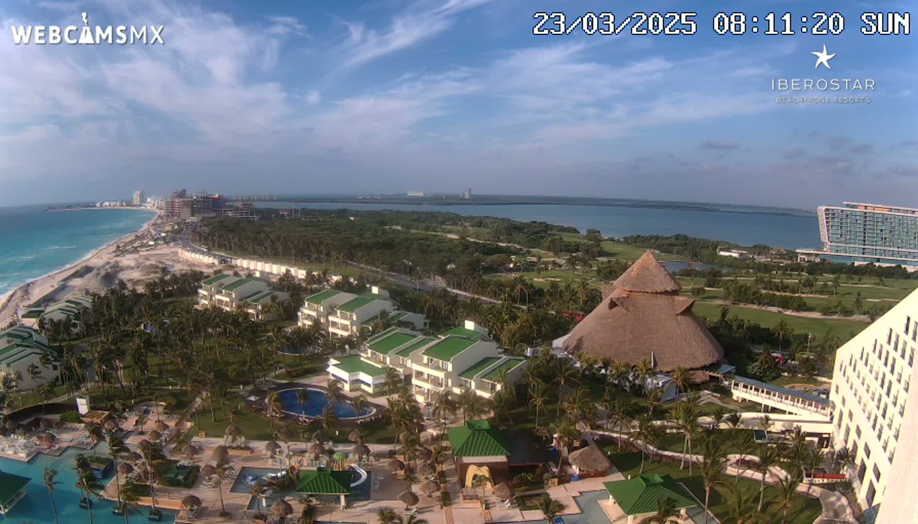 An aerial view shows a resort complex with pools, buildings, and palm trees, situated on a coast next to a body of water under a partly cloudy sky.