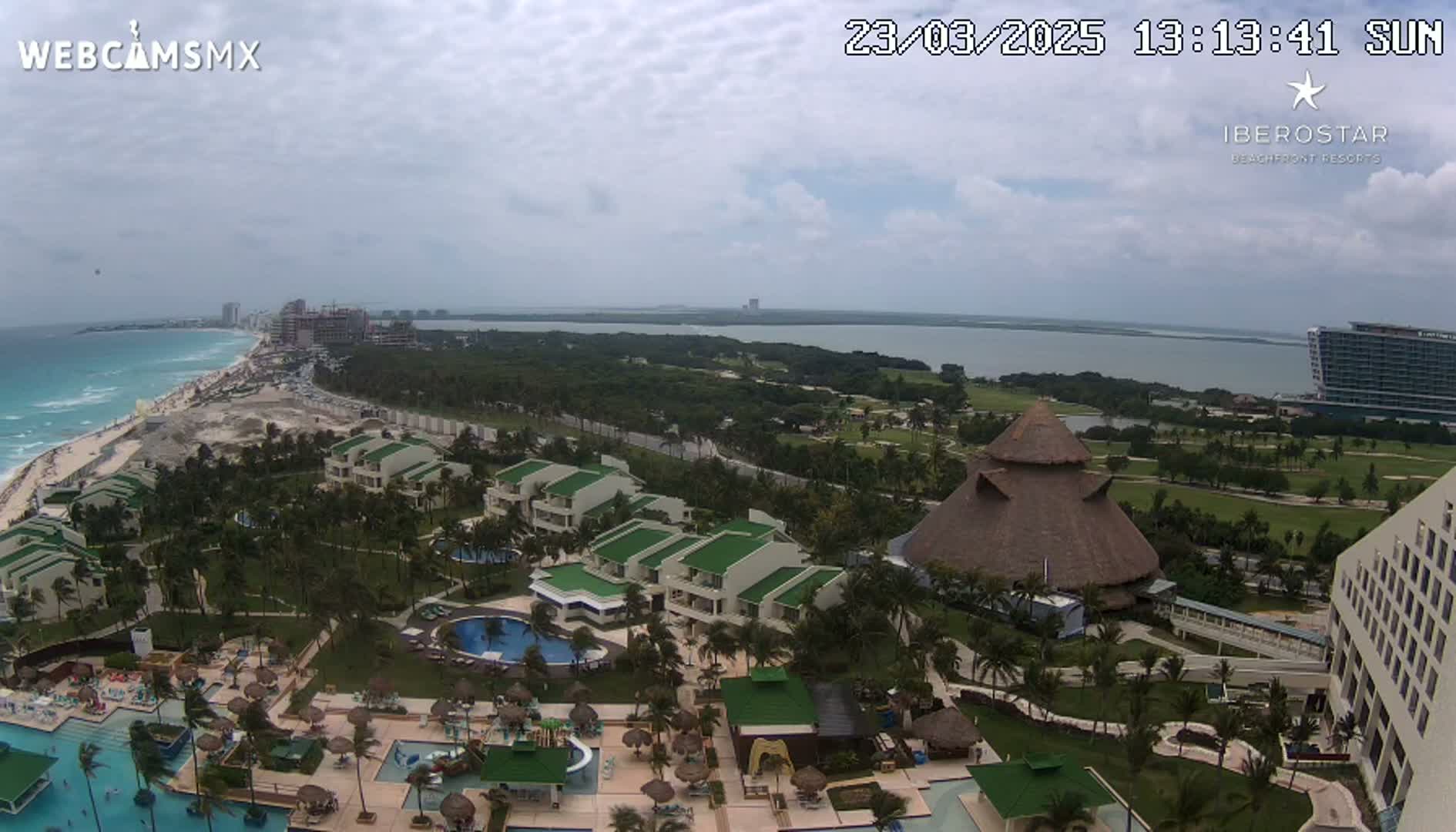 An aerial view shows a resort complex with pools and buildings near a beach and ocean under a partly cloudy sky.
