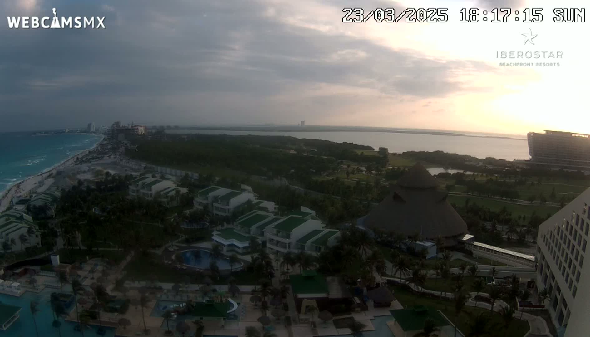 An aerial view shows a resort complex situated on a coastline with a calm ocean under a partly cloudy sunset sky.