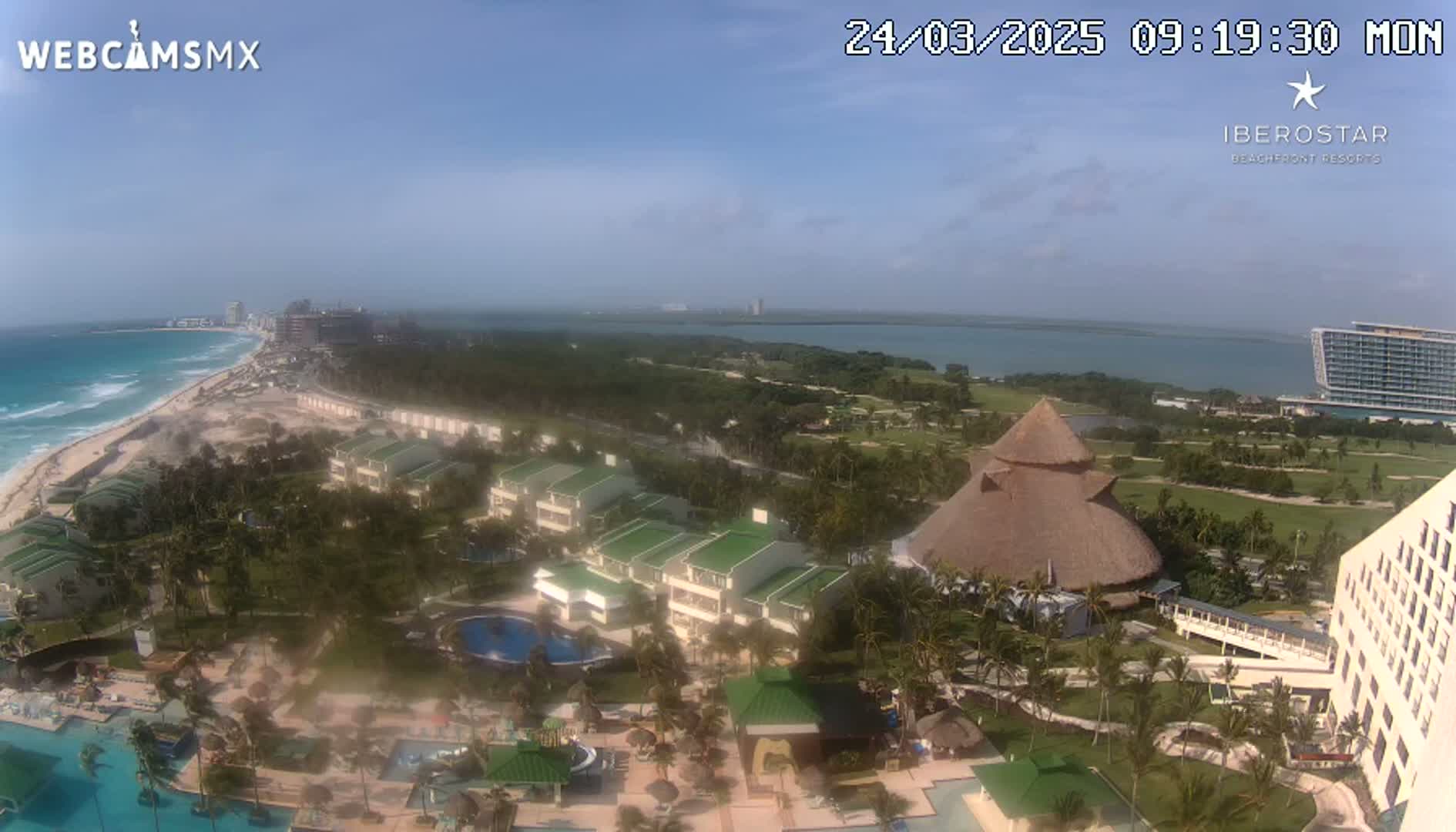 An aerial view of a resort complex on a sunny day, overlooking a beach, ocean, and a golf course.
