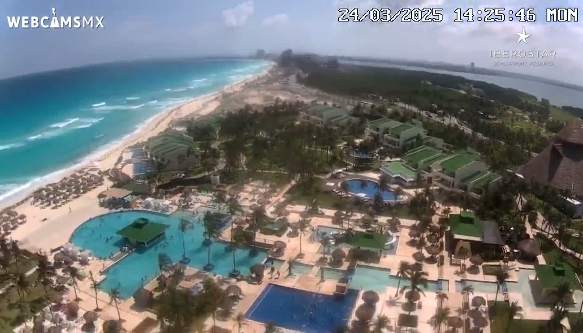 An aerial view shows a resort with numerous pools situated on a beach next to a turquoise ocean under a partly sunny sky.