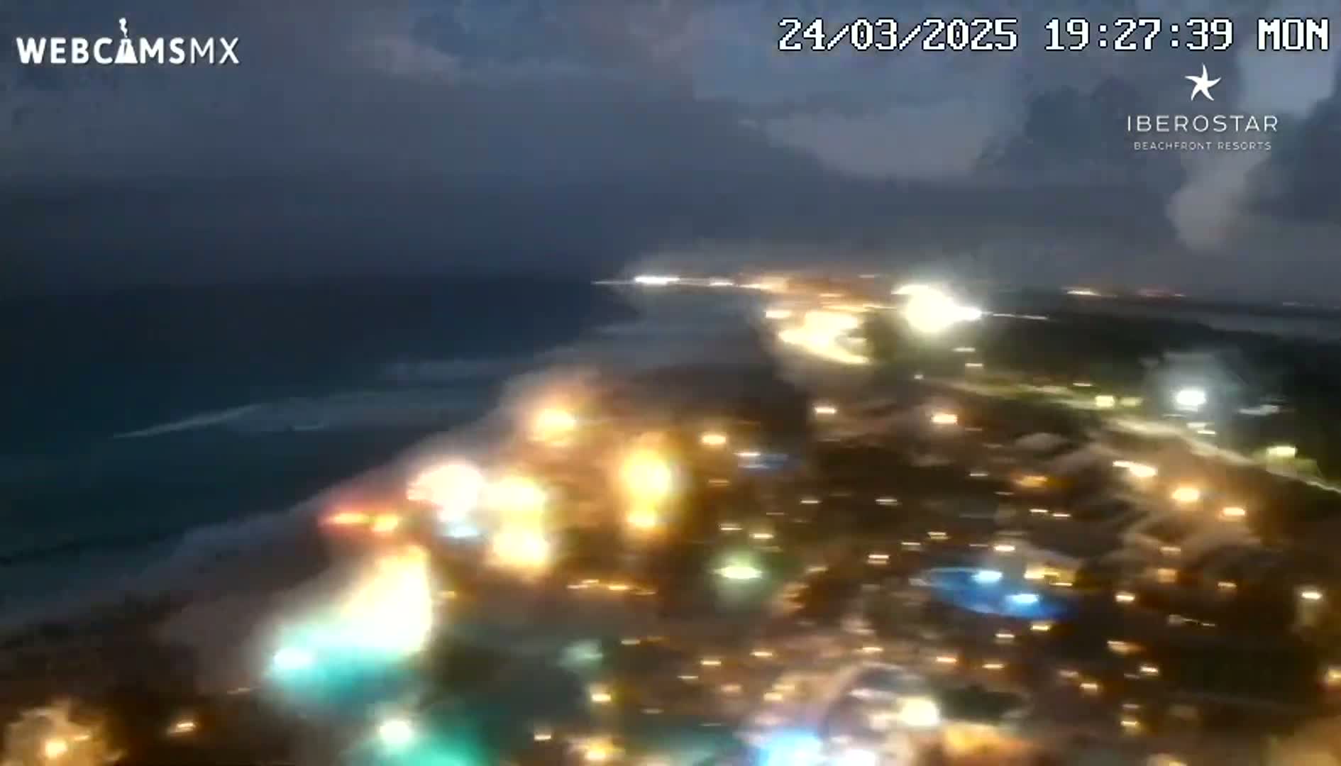 An aerial nighttime view shows a brightly lit resort town next to a dark ocean under a cloudy sky.