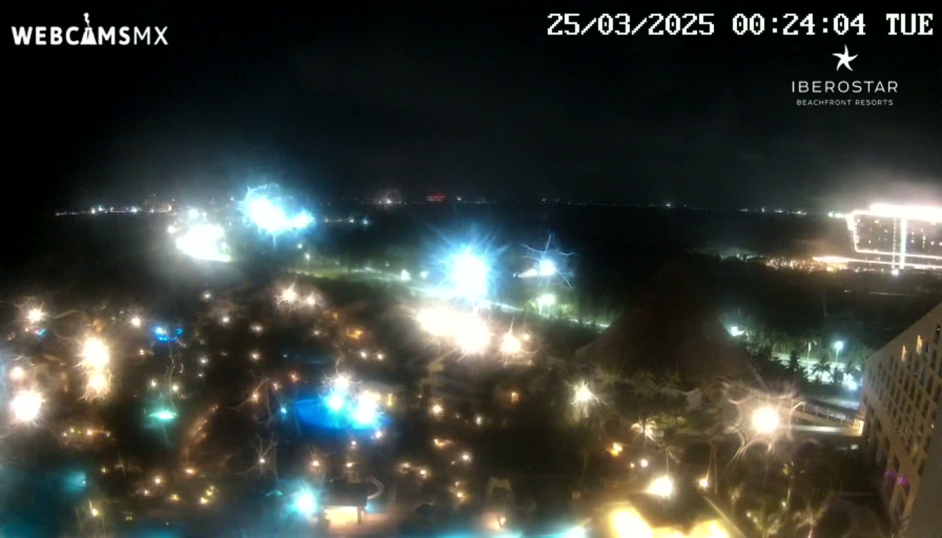 A nighttime aerial view of a resort area shows numerous lights illuminating pools and buildings under a clear, dark sky.