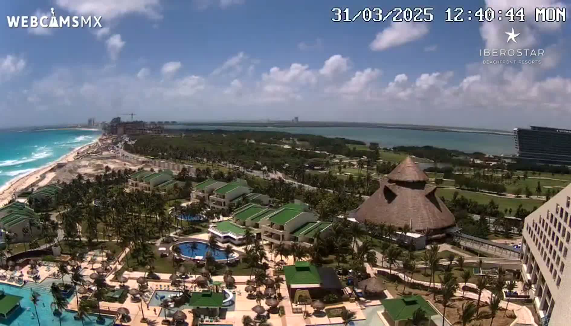 An aerial view shows a resort complex with pools and buildings near a beach and ocean under a partly cloudy sky.