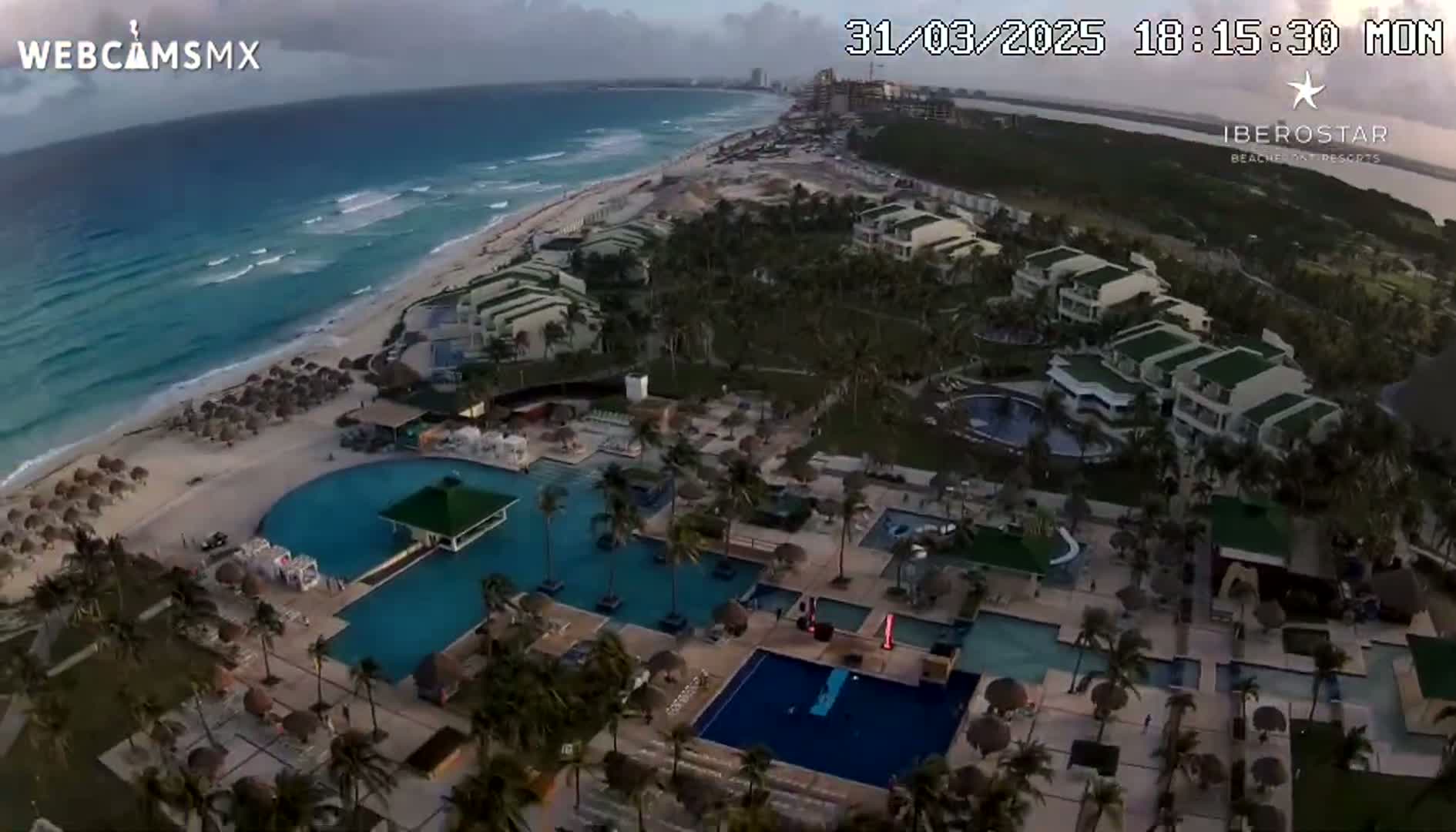 An aerial view shows a resort with multiple pools and buildings next to a beach and ocean under partly cloudy skies.