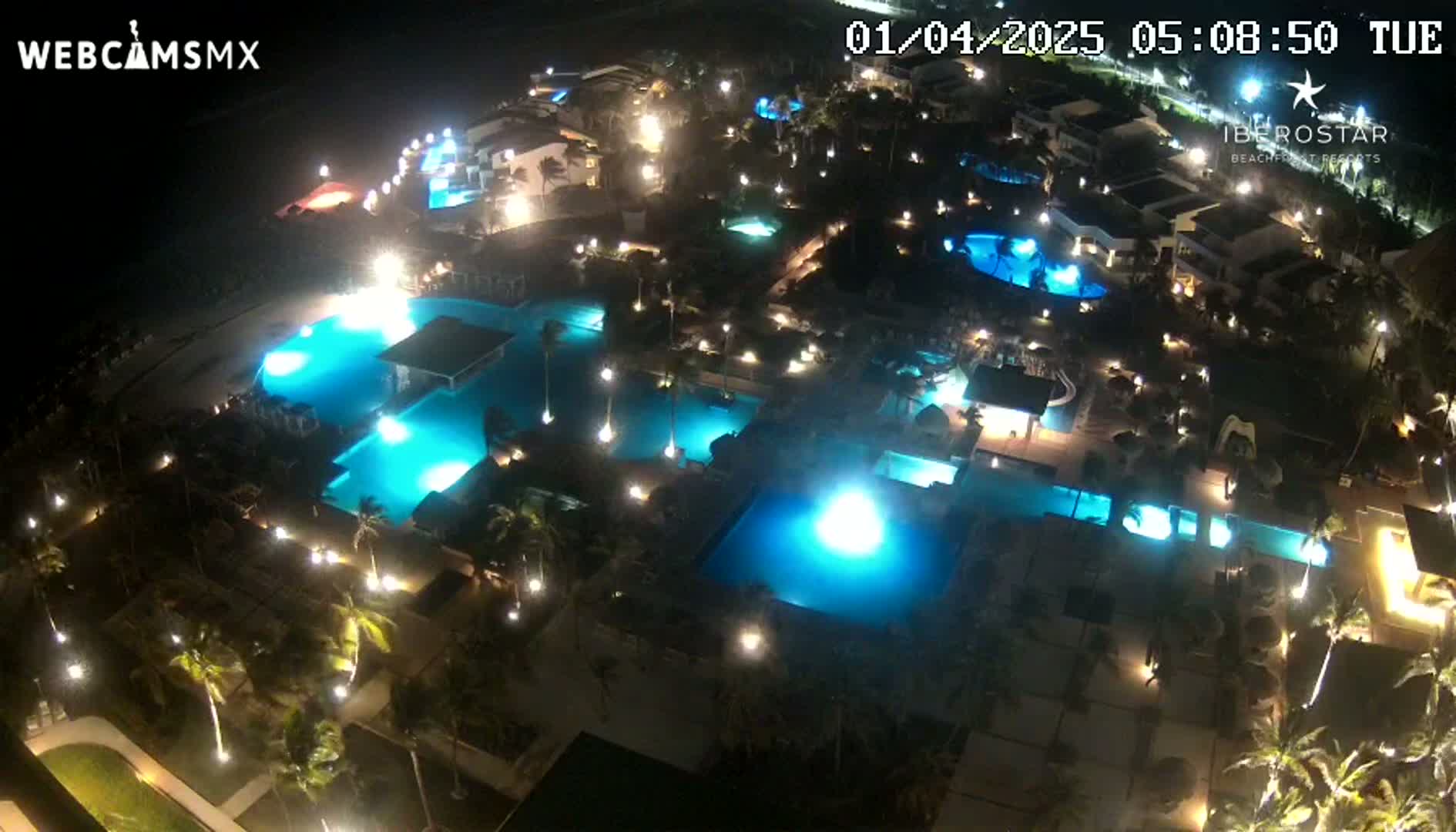 An aerial nighttime view shows a resort with numerous illuminated pools and palm trees surrounding low-rise buildings.