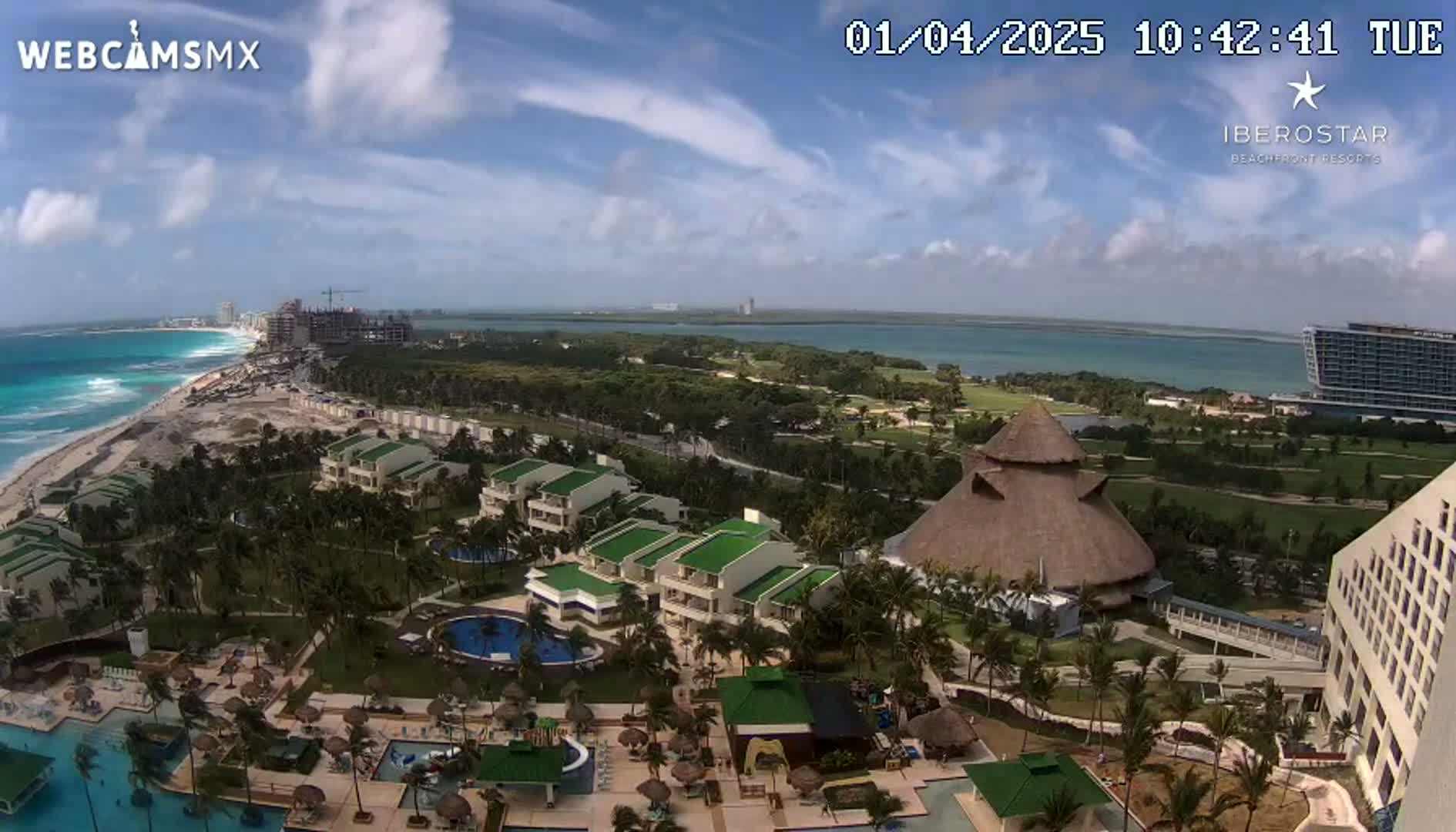 A partly cloudy day overlooks a resort with pools and buildings near a beach and ocean, with other hotels and construction visible in the distance.