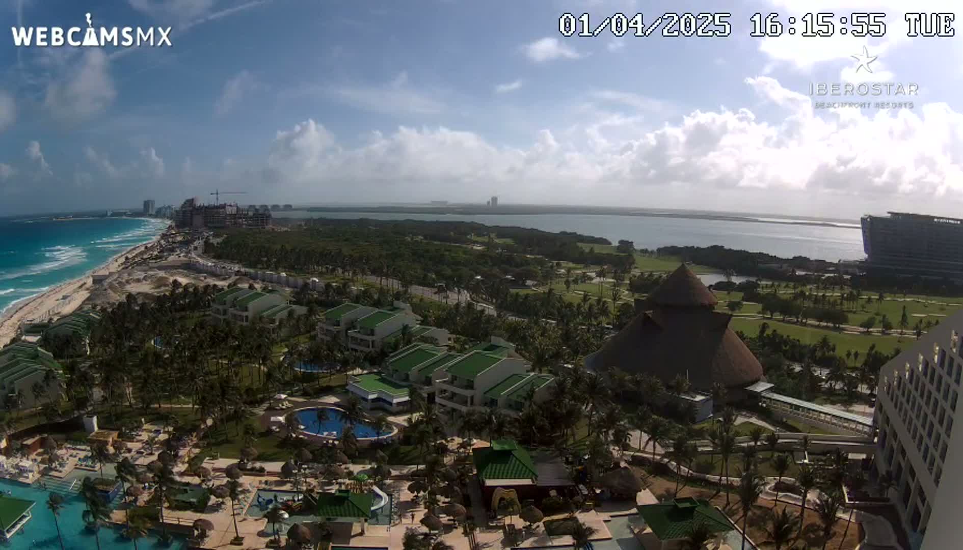 An aerial view of a resort complex situated on a tropical beach, under a partly cloudy sky, shows a turquoise ocean, lush green landscape, and several buildings.