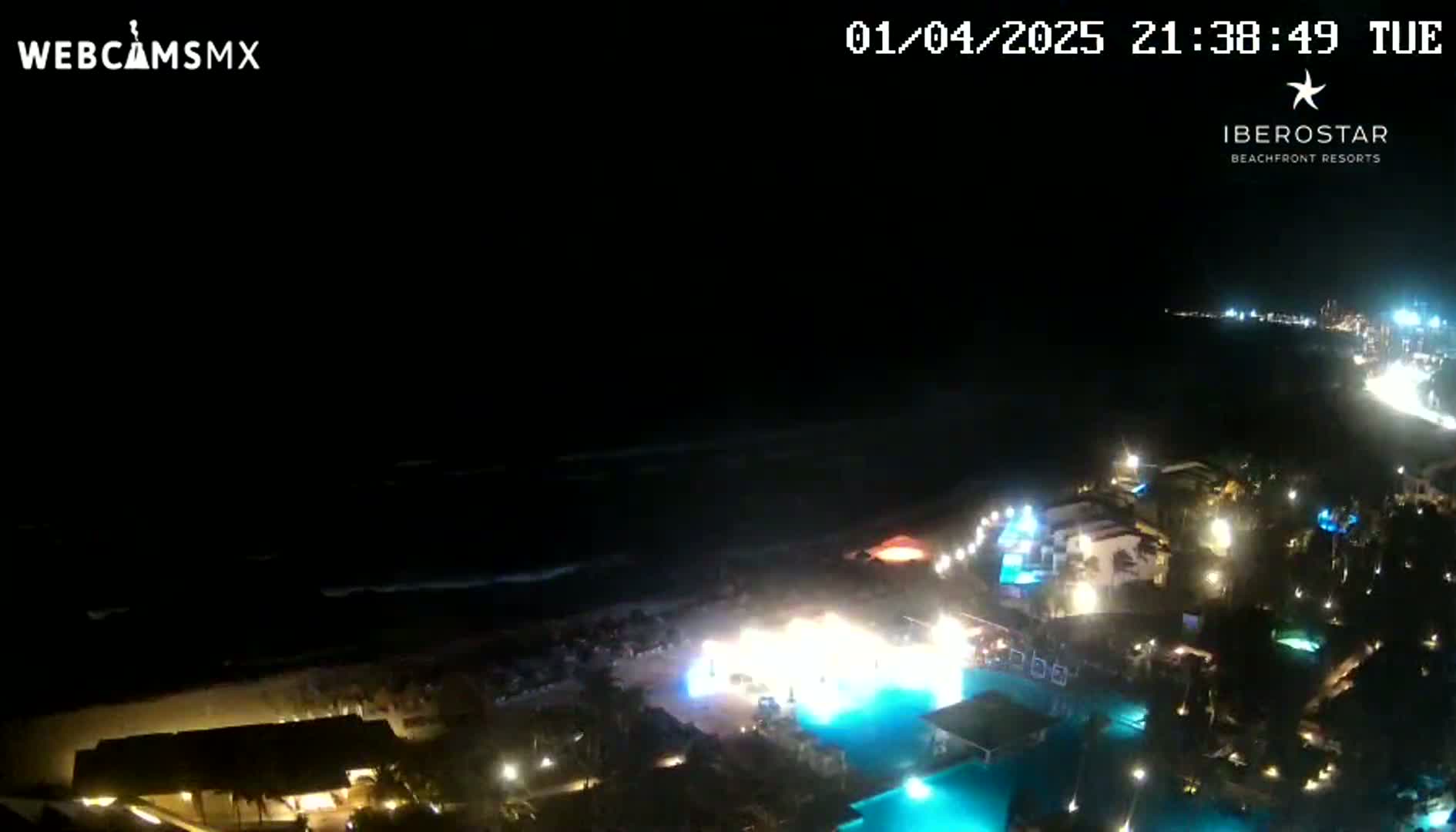 A nighttime aerial view of a resort on the coast, showing a dark ocean, lit-up buildings and pools, and a bright spectacle on the beach.