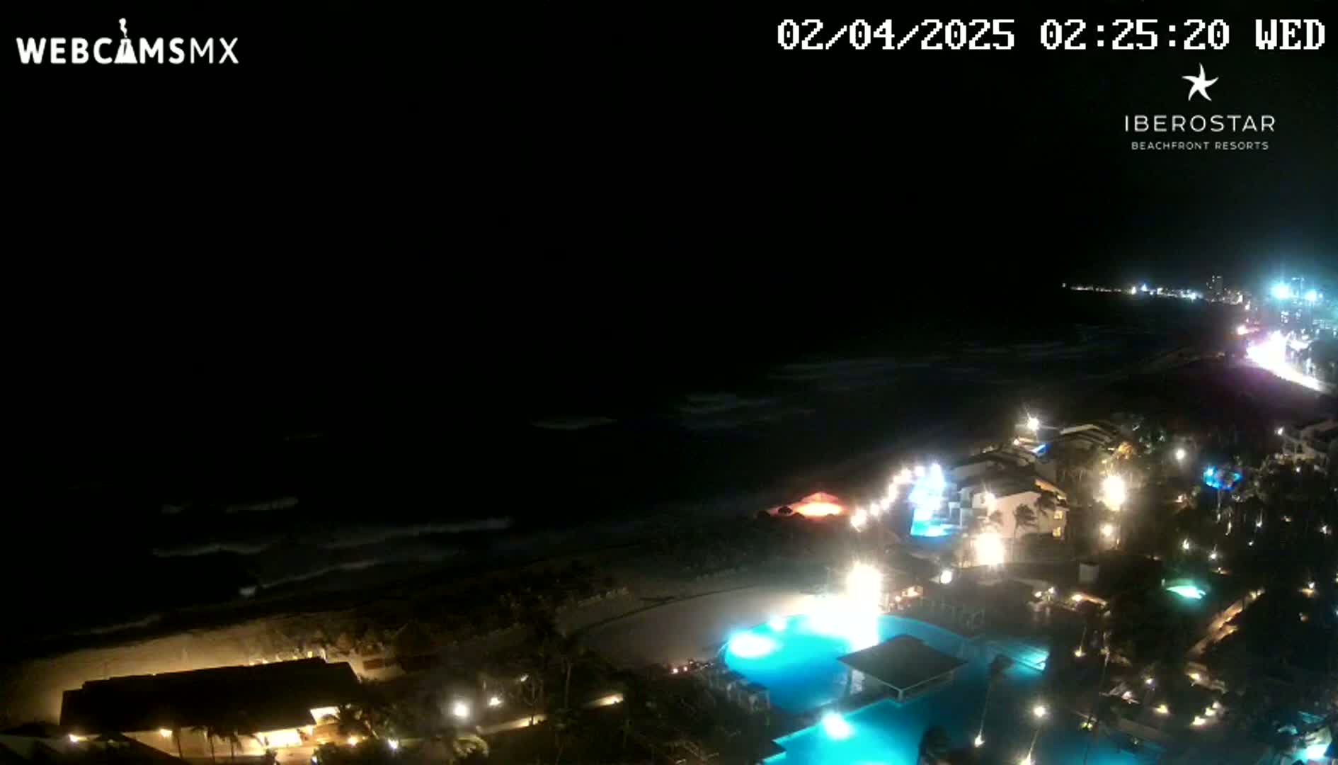 A nighttime aerial view shows a resort with illuminated pools and buildings along a dark beach with gently waving ocean under a clear, dark sky.