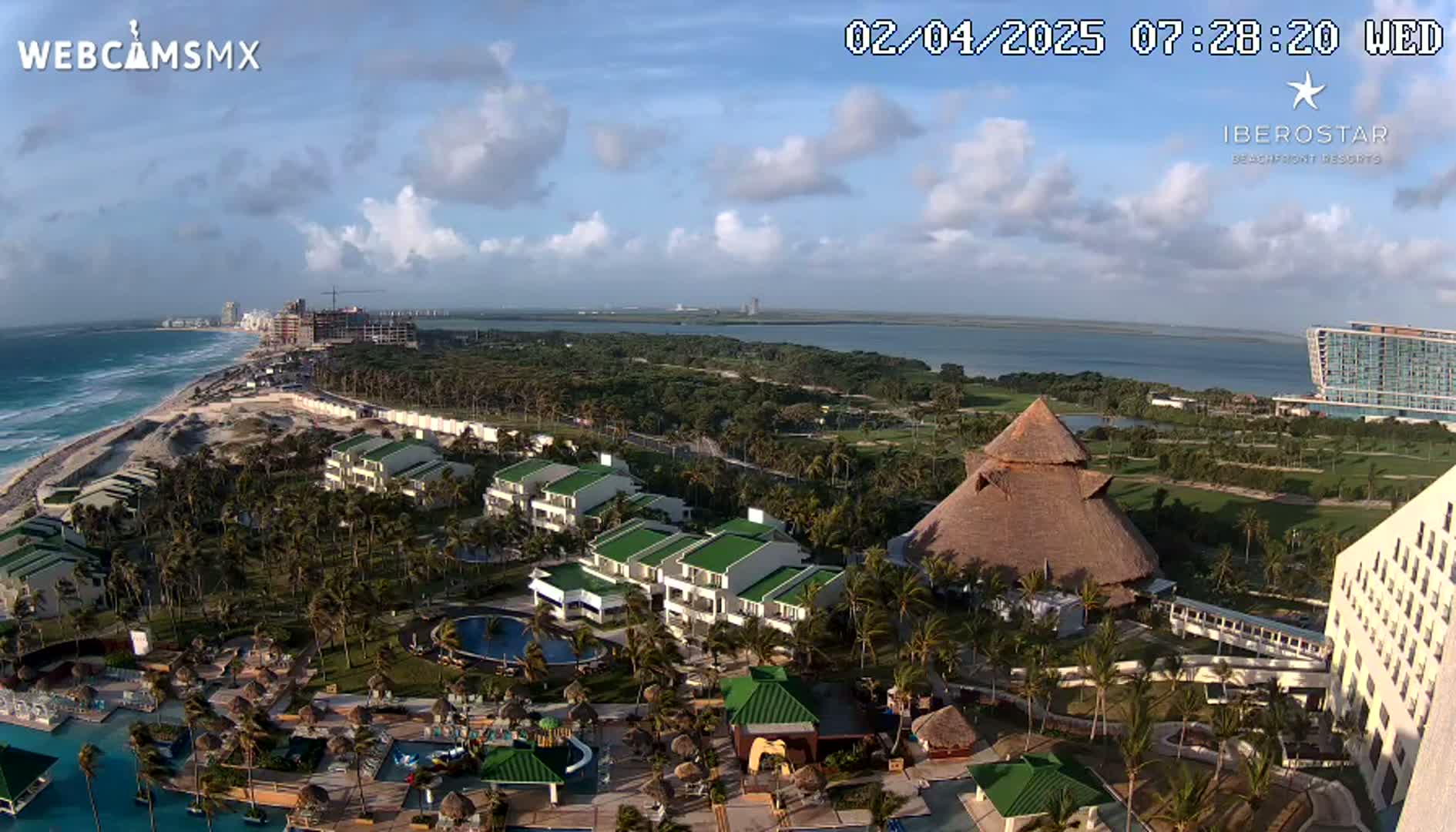An aerial view of a tropical resort, showing several buildings, pools, and palm trees, under partly cloudy skies and next to a beach.