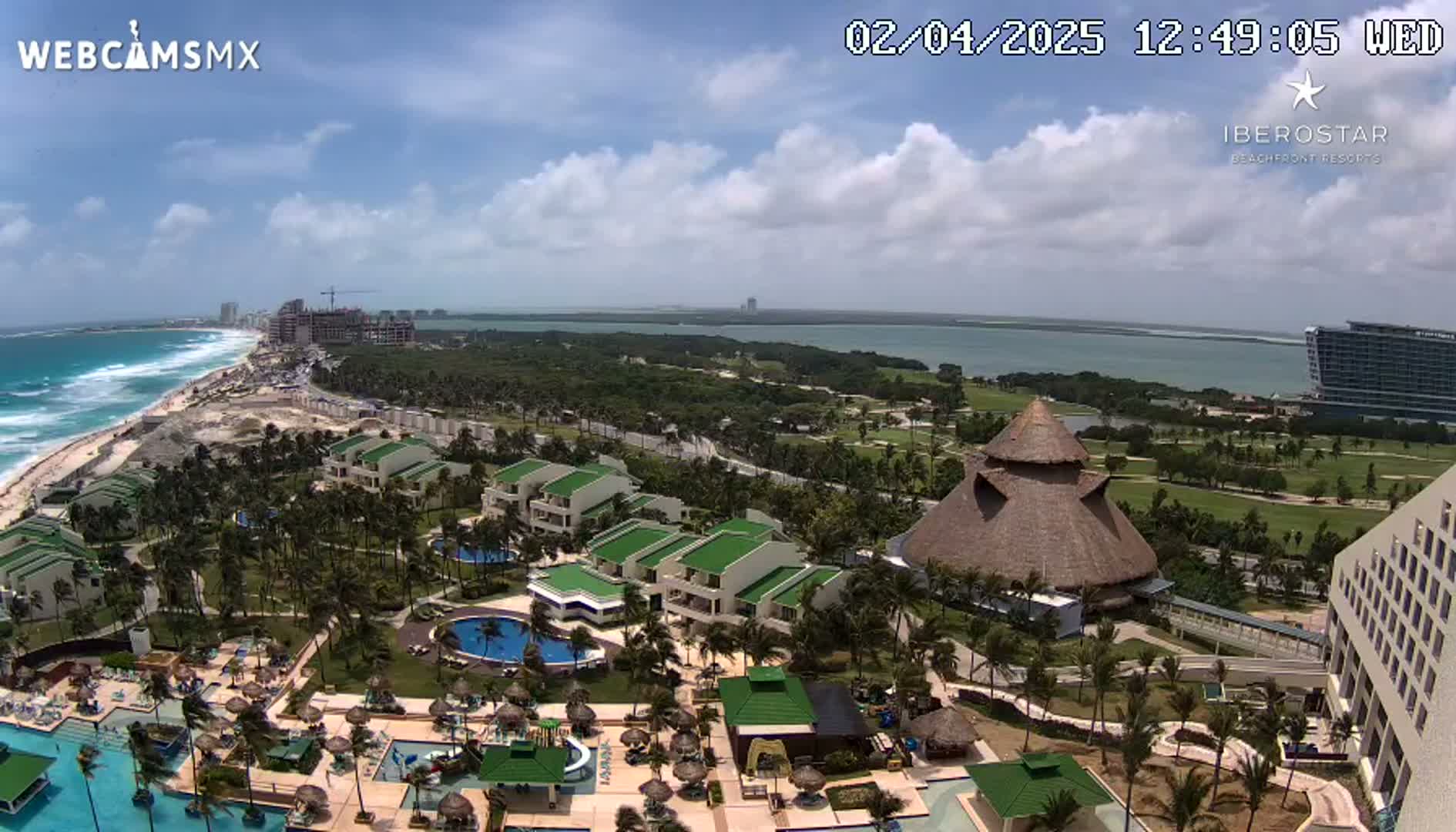 An aerial view shows a resort complex with pools, buildings, and lush landscaping next to a beach with waves under a partly cloudy sky.