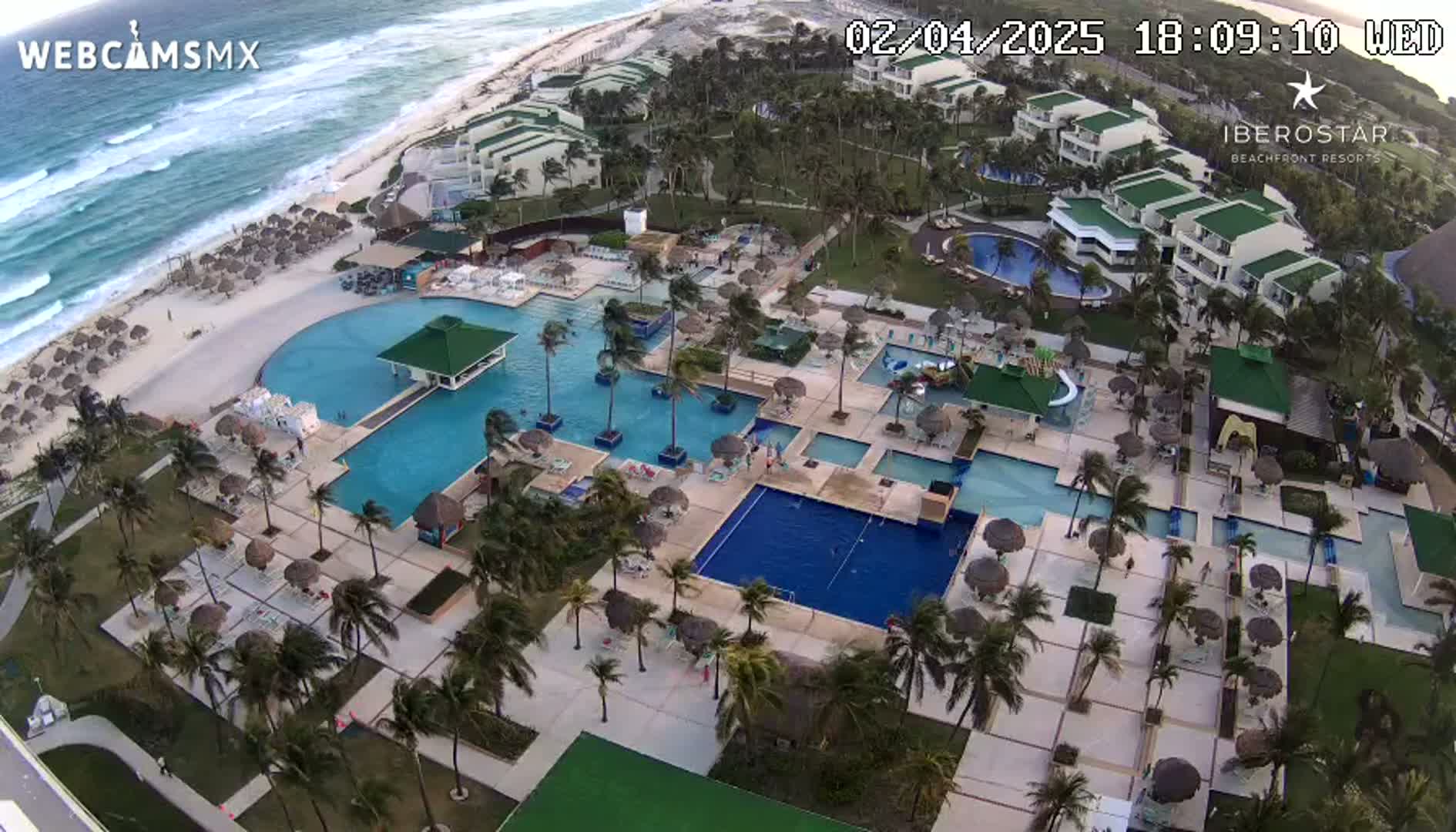 An aerial view shows a resort with multiple pools, surrounding palm trees, and a beach with wavy ocean under a somewhat windy sky.