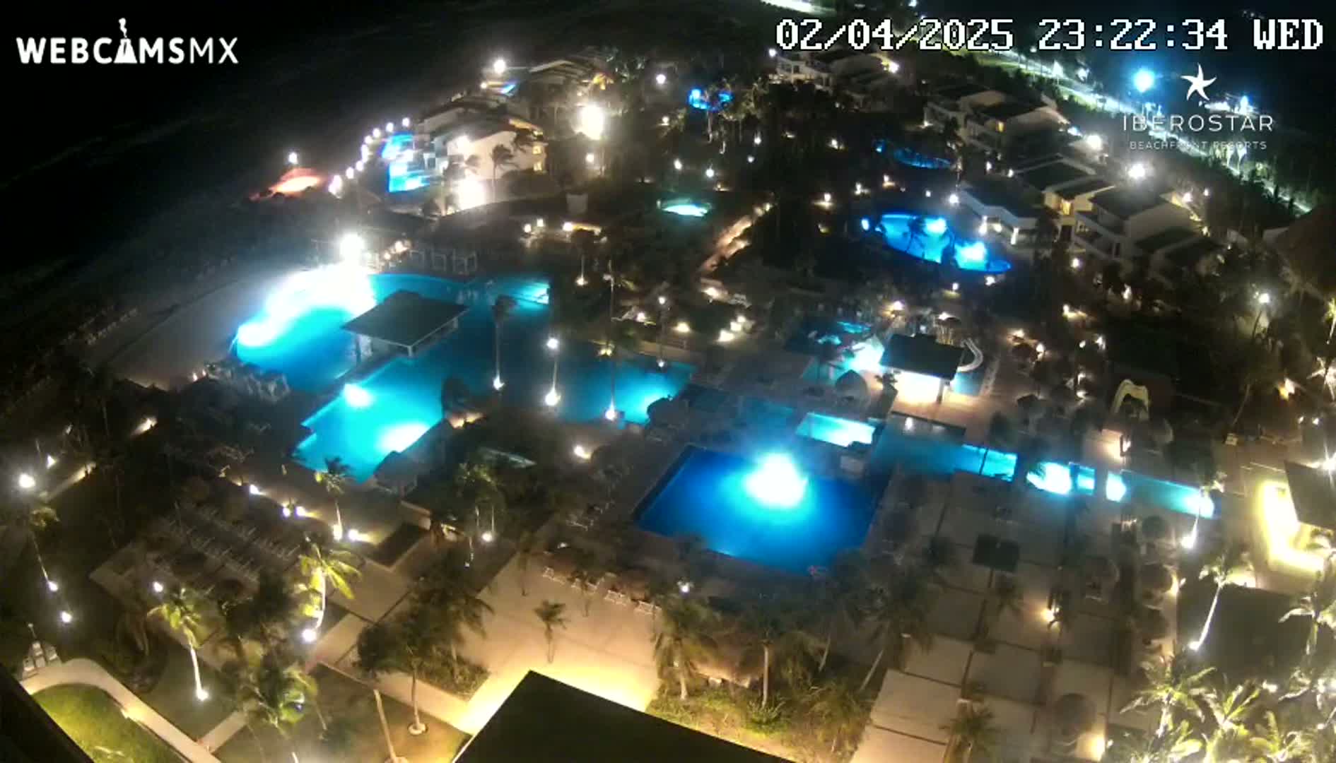 An illuminated resort at night, featuring numerous pools and palm trees, is seen from an elevated vantage point.