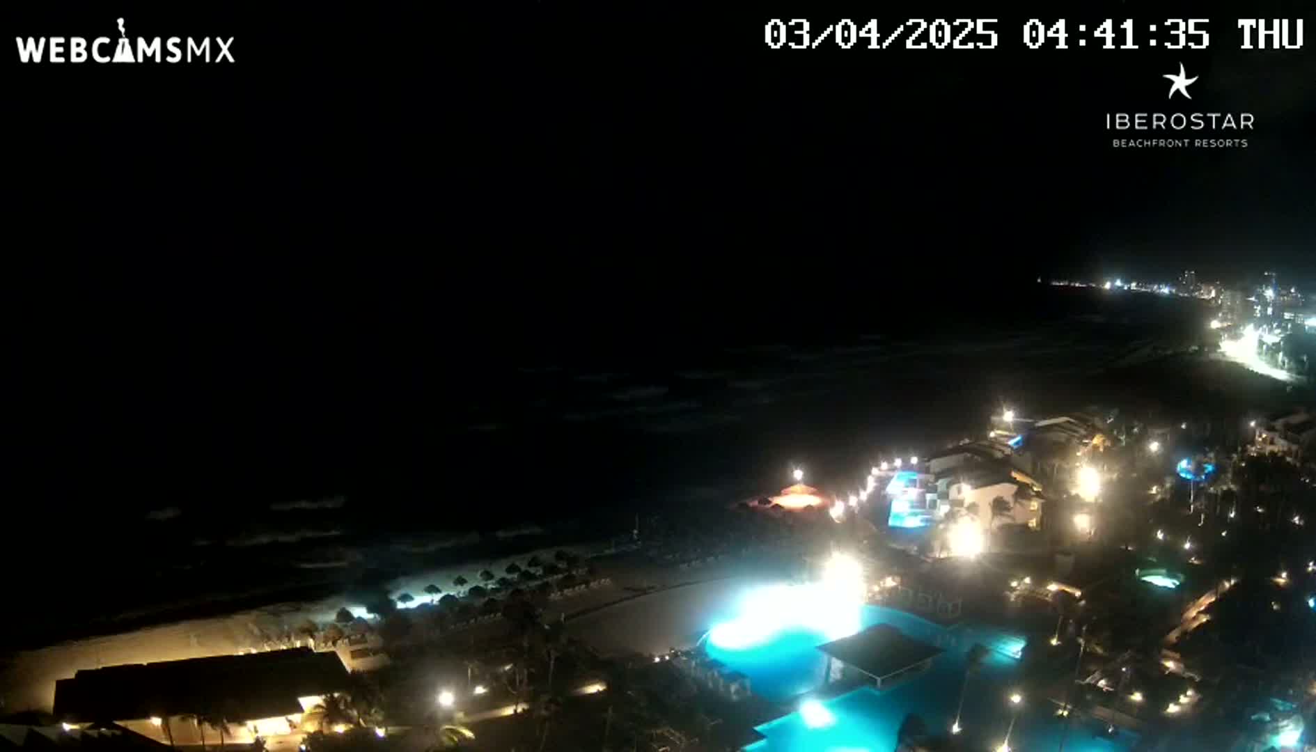 A nighttime, aerial view shows a resort area along a dark ocean shoreline with lit buildings, pools, and palm trees under a clear, dark sky.