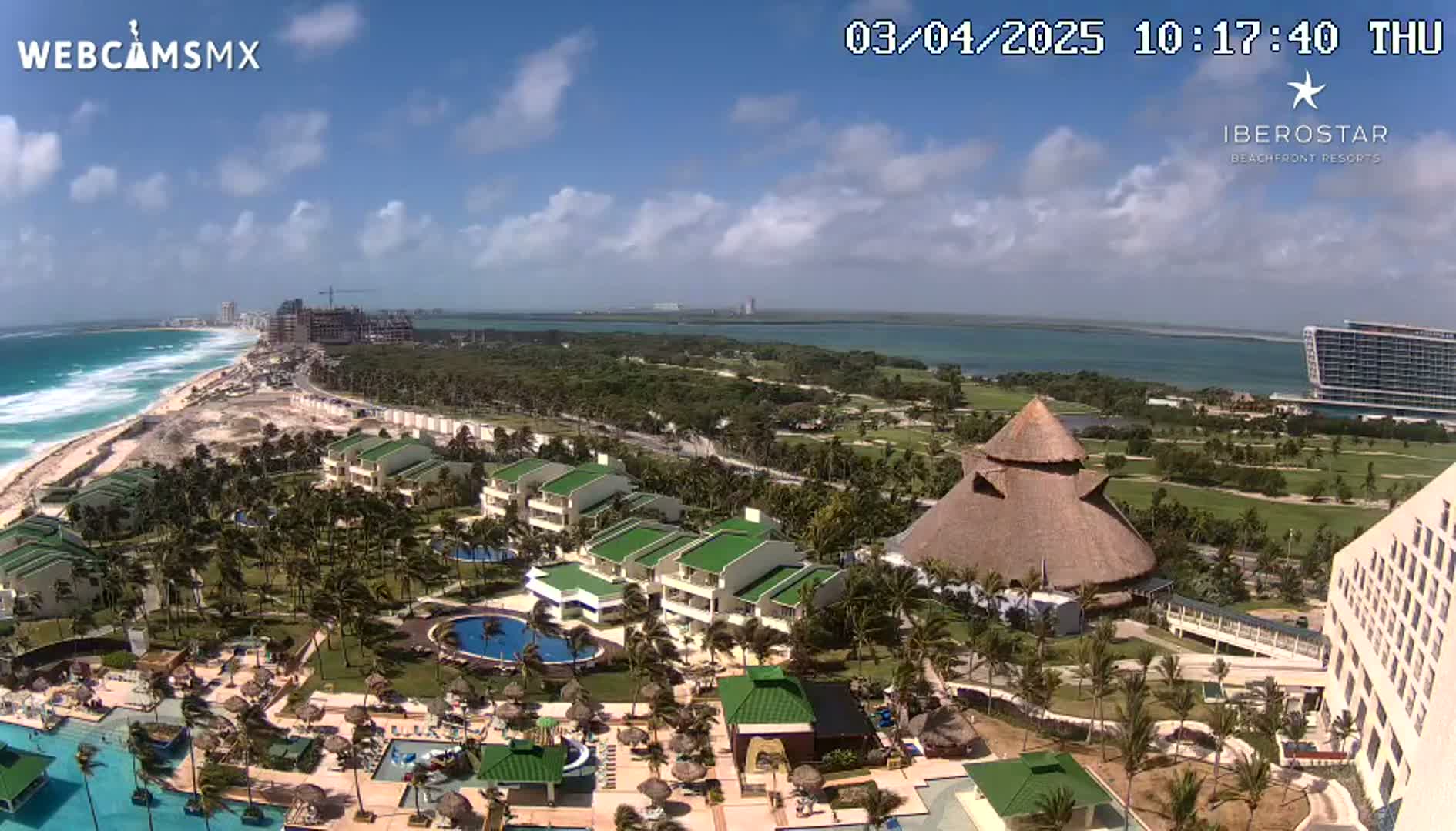 An aerial view shows a resort complex with pools, palm trees, and buildings near a beach with waves under a partly cloudy sky.
