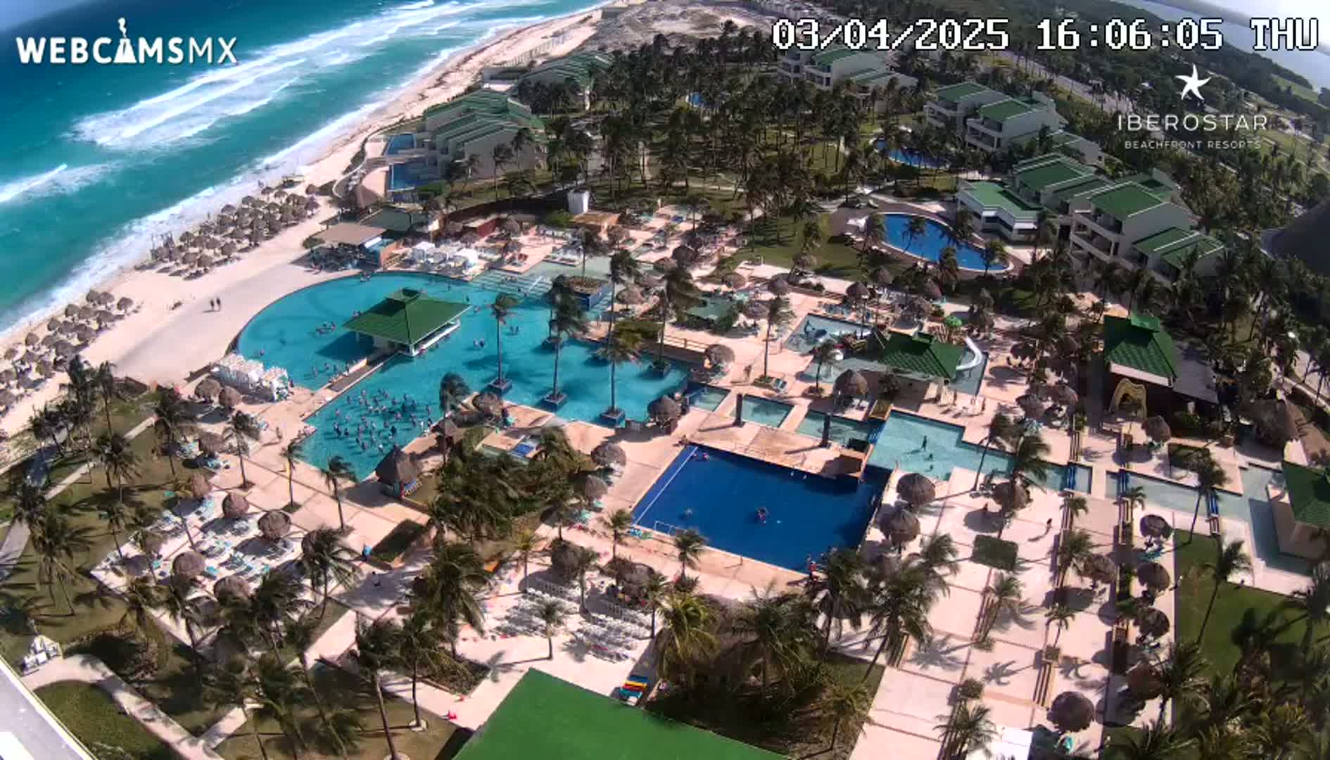 An aerial view shows a resort with multiple pools, a beach, and palm trees on a sunny day with wavy turquoise water.