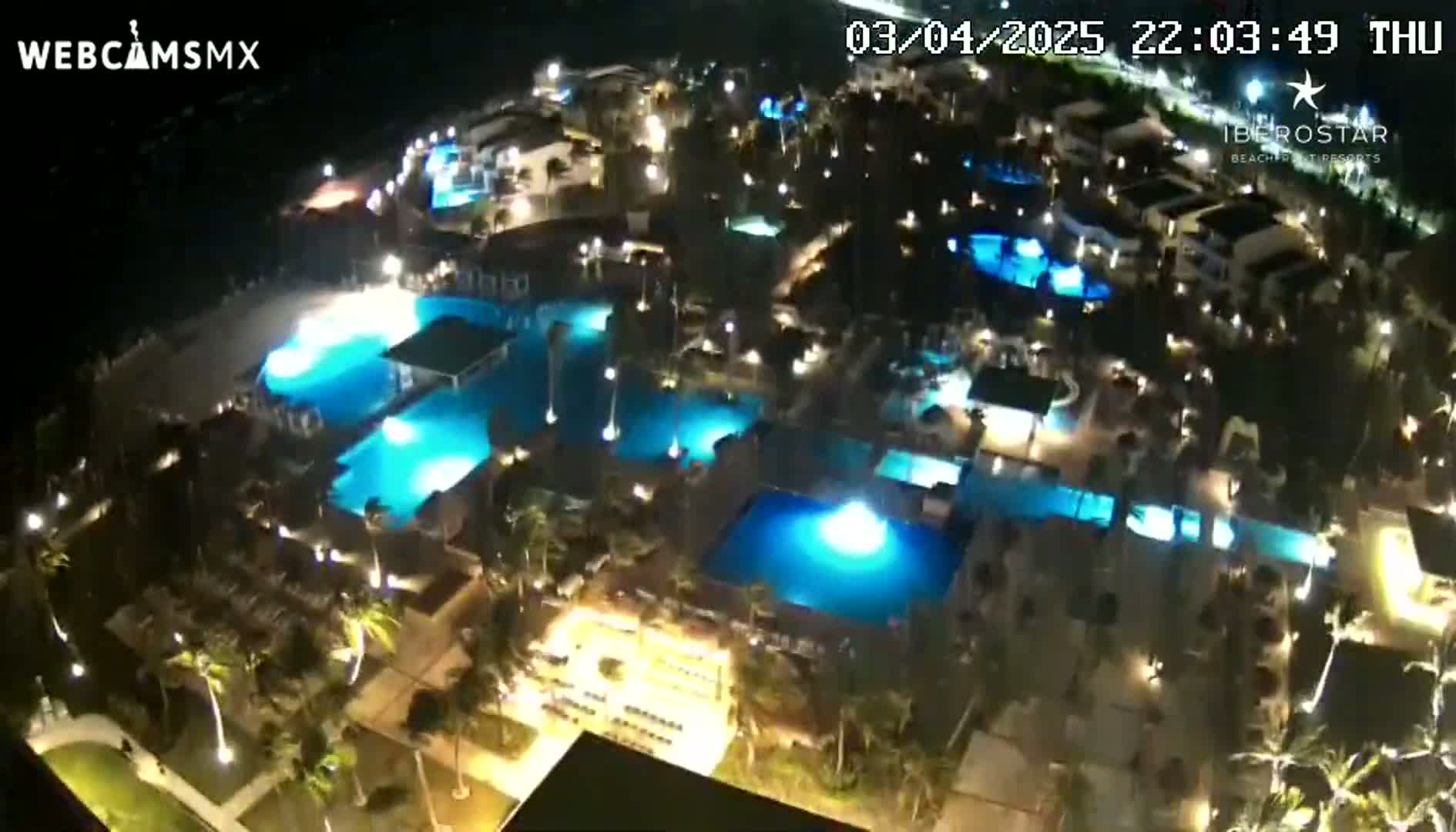 An aerial nighttime view shows a resort complex with numerous illuminated swimming pools surrounded by buildings and palm trees.