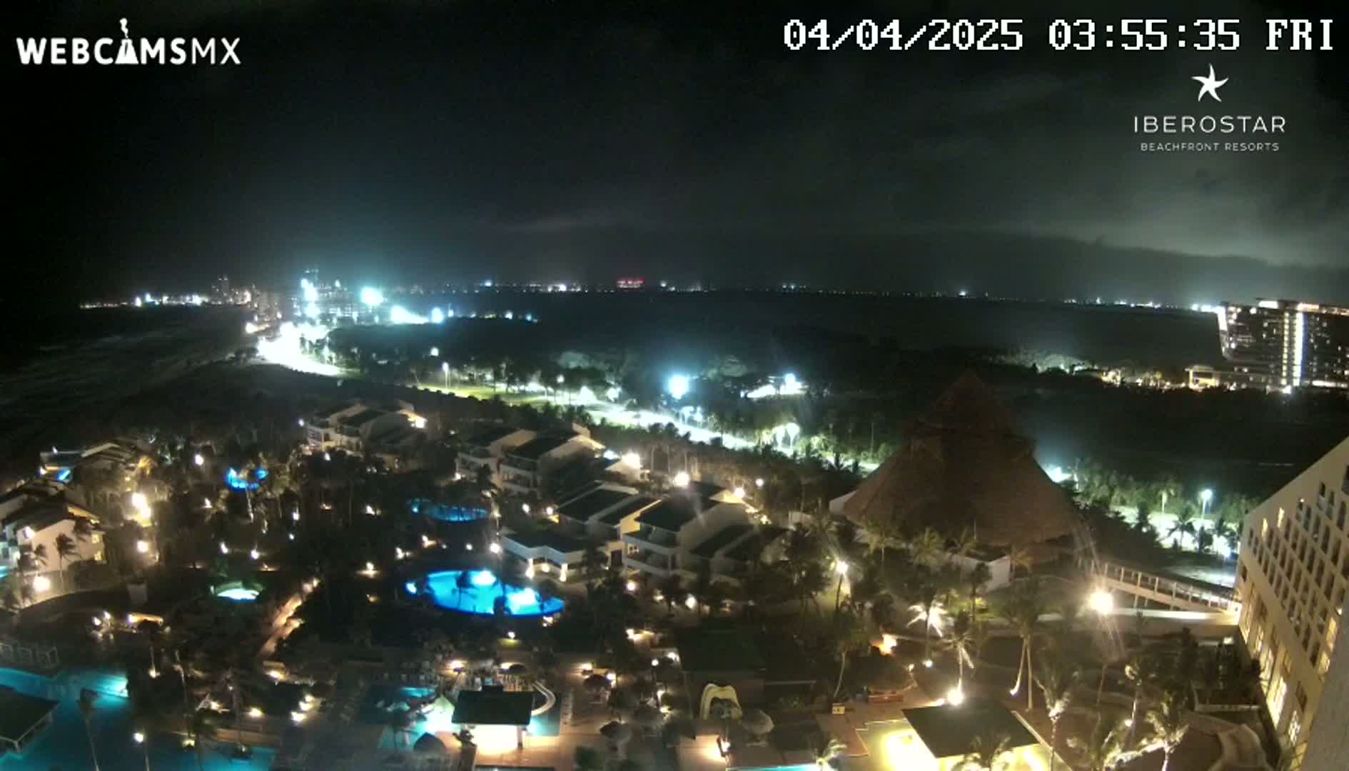 A nighttime aerial view of a resort complex with illuminated buildings, pools, and landscaping, overlooking a dark ocean under a cloudy sky.