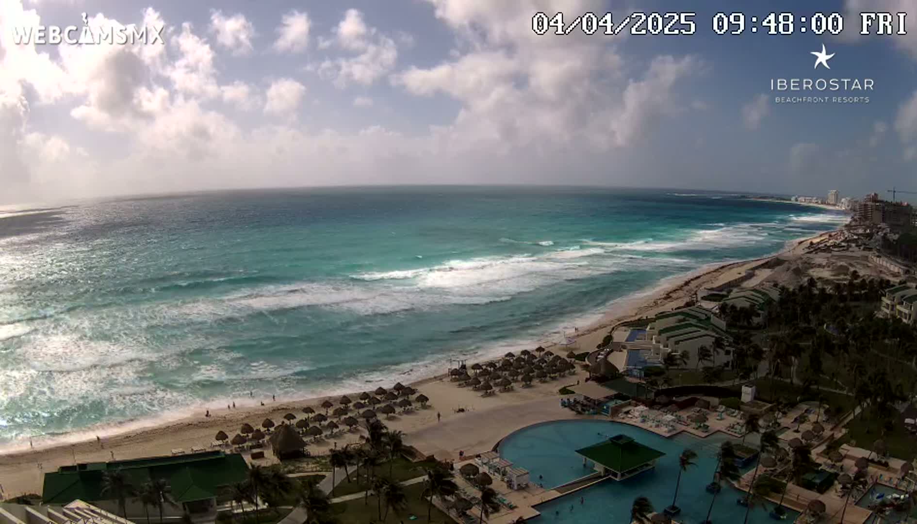 A partly cloudy day shows turquoise ocean waves breaking on a sandy beach with resort buildings, pools, and beach umbrellas.