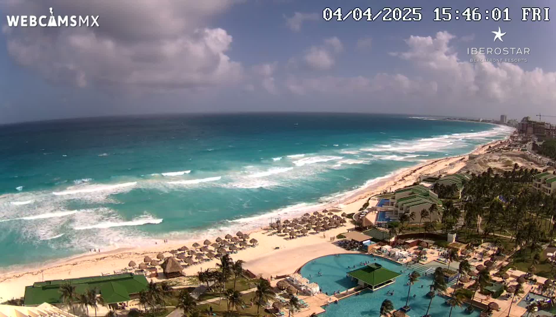 A partly cloudy day shows a beach with turquoise water and moderate waves, along with a resort featuring pools and palm trees.