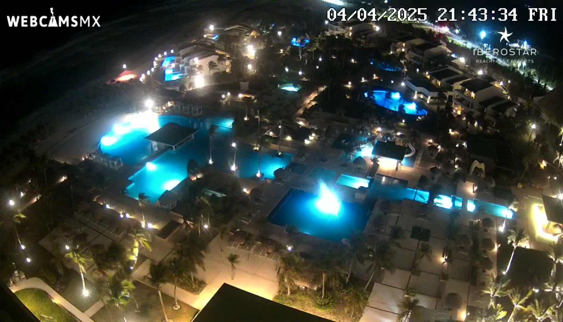 An illuminated resort at night, featuring multiple pools and surrounding buildings, is seen from an elevated perspective.