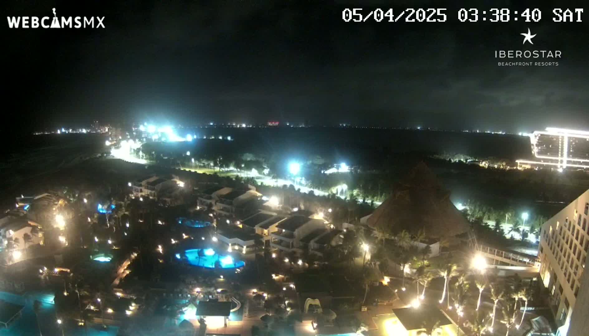 A nighttime aerial view of a resort complex with pools and palm trees, situated near a coastline under a partly cloudy sky.