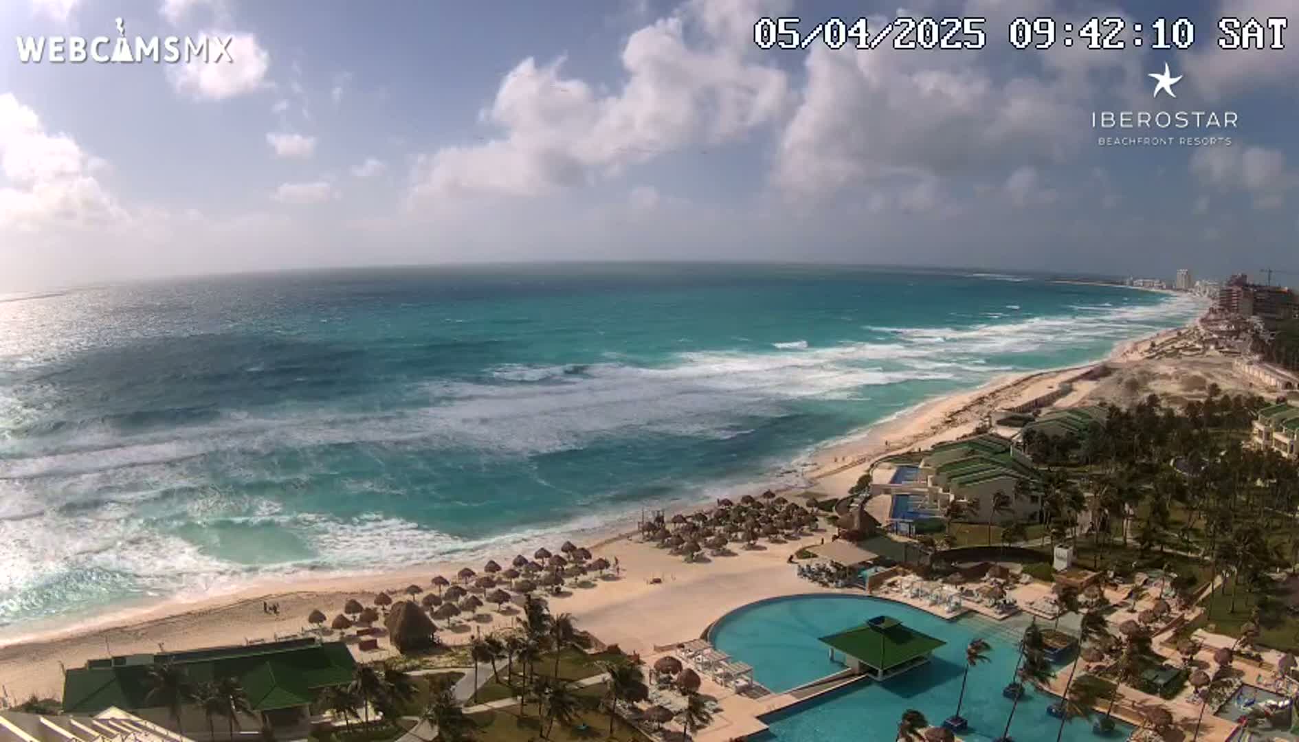 A beach resort with a large swimming pool overlooks a partly cloudy sky and a turquoise ocean with moderate waves.