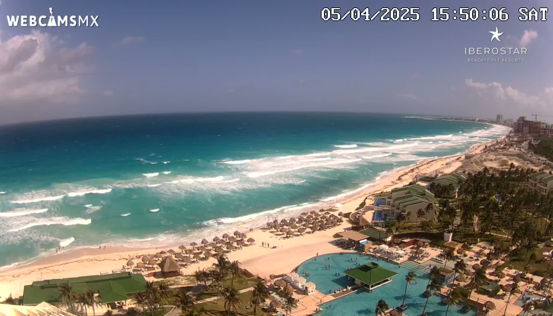 An aerial view shows a resort beach with turquoise water, white sand, and palm trees under a partly cloudy sky with moderate waves.