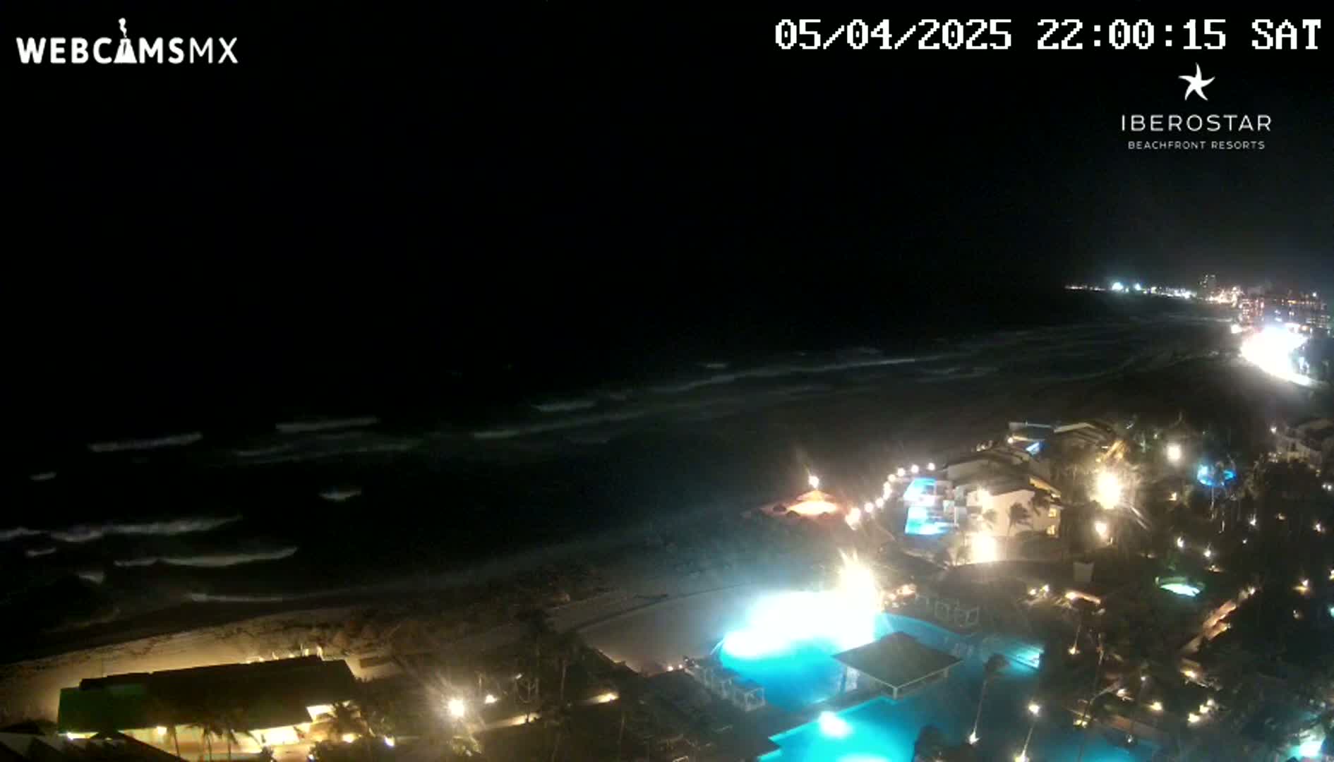 A nighttime view shows a resort coastline with illuminated pools and buildings overlooking a dark ocean with moderate waves.