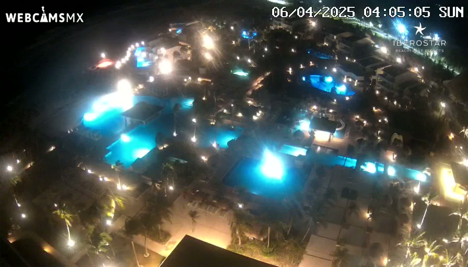 An aerial nighttime view of a resort complex with numerous illuminated swimming pools surrounded by palm trees and buildings.