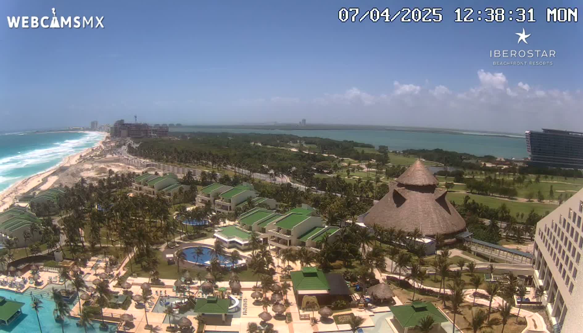 An aerial view of a resort on a sunny day, showing a beachfront property with pools, buildings, and lush landscaping, overlooking a calm ocean and a lagoon.